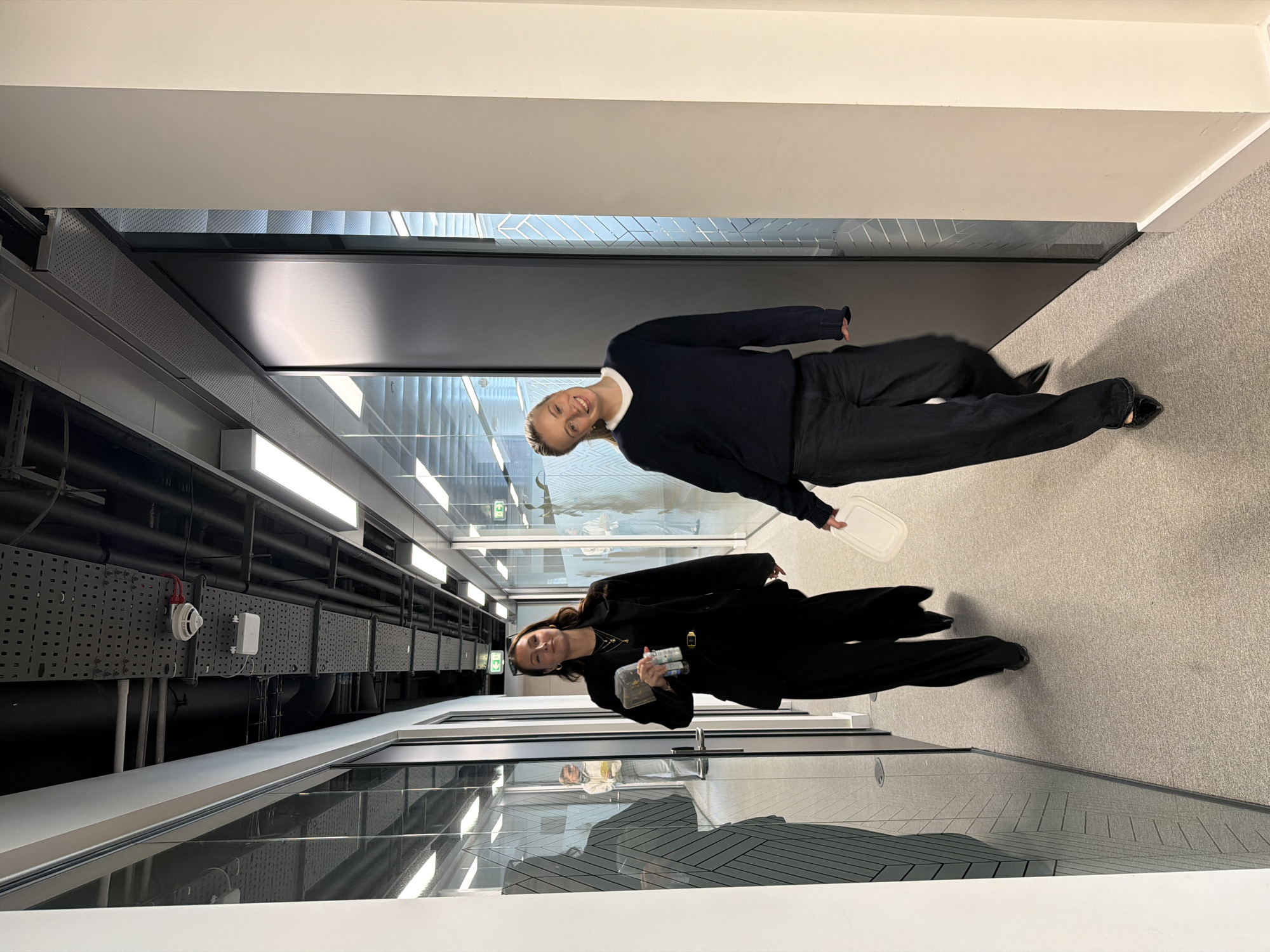 Two women stand in a brightly lit office hallway. The hallway is flanked by large glass windows on one side and a solid wall on the other. The floor is carpeted in a neutral grey. Overhead, black pipes and ventilation systems are visible. One woman wears a dark blazer and pants. The other woman wears a dark sweater and pants. The location appears to be an office building interior, potentially in London, United Kingdom, given the architectural style often found in commercial spaces there.