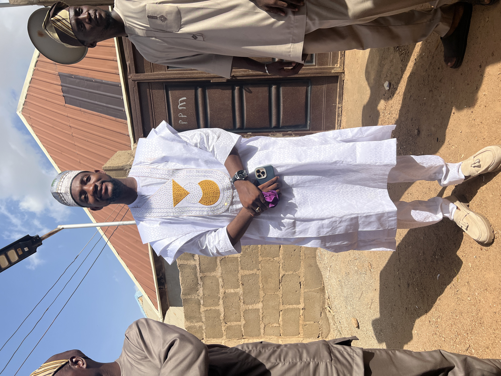 Two men are standing outdoors in Rigasa, Nigeria. One man is wearing a white, patterned robe with gold embroidery, a white cap, and beige shoes. He is holding a smartphone and a purple object. The other man is wearing a tan outfit with a matching hat and black shoes. Behind them are a building with a brown door and a red roof, a brick wall, and a dirt ground. A streetlight is visible in the background.