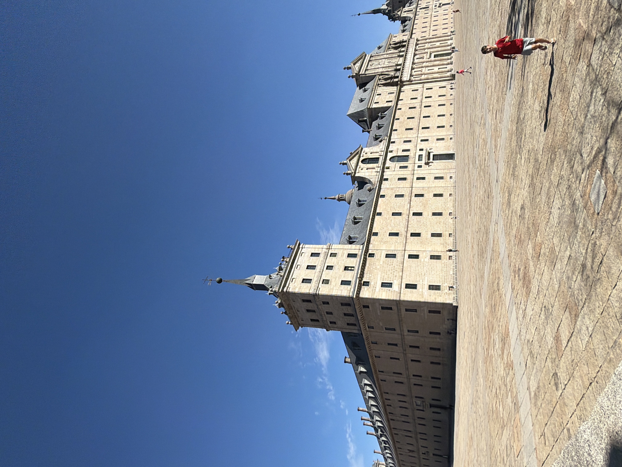 The image depicts a wide shot of the Plaza de la Virgen de la Herrería in San Lorenzo de El Escorial, Spain, featuring the Monastery of El Escorial as the dominant background element. The monastery, a vast Renaissance structure, is characterized by its imposing stone facade, numerous windows, and a prominent central tower topped with a spire and a cross. The sky above is a clear, vibrant blue with a few wispy clouds. The plaza itself is paved with large, rectangular stone slabs, showing variations in color and texture, with some lighter rectangular sections.

In the foreground and midground of the plaza, several people are present. On the right side of the image, a young boy wearing a red t-shirt and grey shorts is running towards the viewer, casting a long shadow on the stone pavement. Further into the plaza, other individuals, including children, are visible as small figures, some walking or running, creating a sense of scale against the massive architecture. The scene is brightly lit by sunlight, suggesting it is daytime, and the overall atmosphere is one of open space and activity within a historical setting.