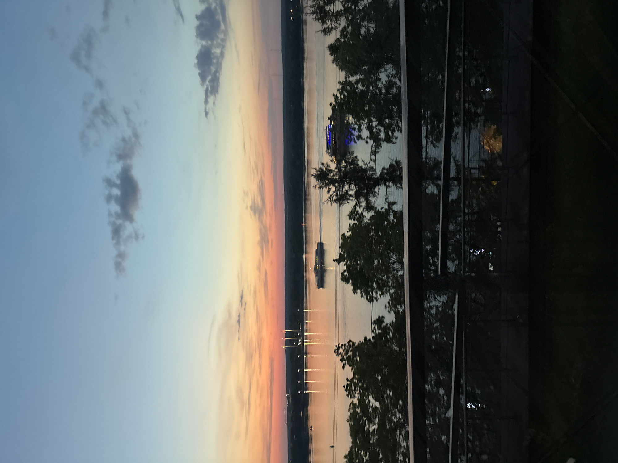 The image shows a sunset over the Kama River in Perm, Russia. The sky is colored with gradients of blue, orange, and pink. Silhouetted against the sunset are trees in the foreground and the opposite bank of the river. Two vessels are visible on the river. The lights of the city of Perm are visible along the riverbank, reflected in the water. A railing and the silhouette of a building are present in the lower right corner of the image.