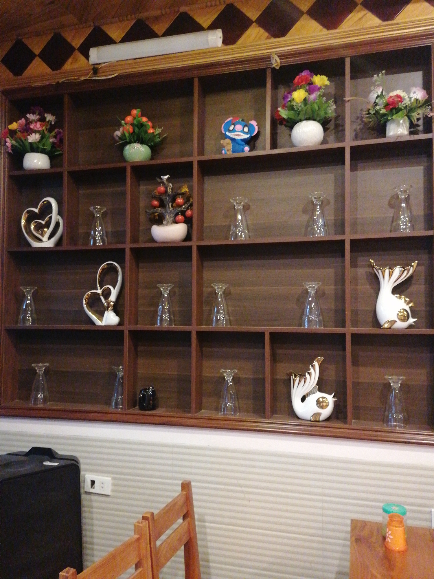 The image shows a dark wooden display cabinet with multiple shelves, each containing various decorative items such as flowers, ceramic figures, and glassware. The cabinet is situated against a wall covered with horizontal striped wallpaper. Below the cabinet, there is a wooden chair and part of a black suitcase, along with a wooden table with an orange and green container. The location is identifiable as Sa Pa Ward, Vietnam.