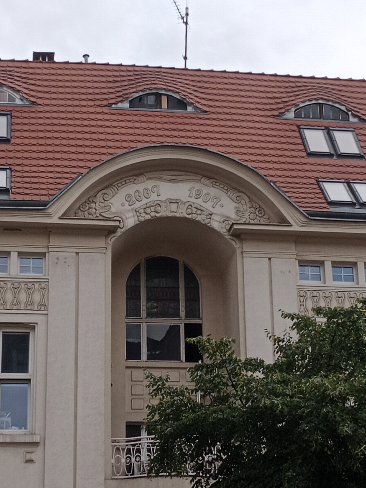 The image shows the facade of a building with a red tiled roof. There are dormer windows on the roof and several regular windows on the facade. An arched decorative element above a large window contains the inscriptions "2007" and "1907". A tree with green foliage obscures part of the lower section of the building and the balcony. The sky is overcast. The building is located at 14 Gdańska Street in Bydgoszcz, Poland.
