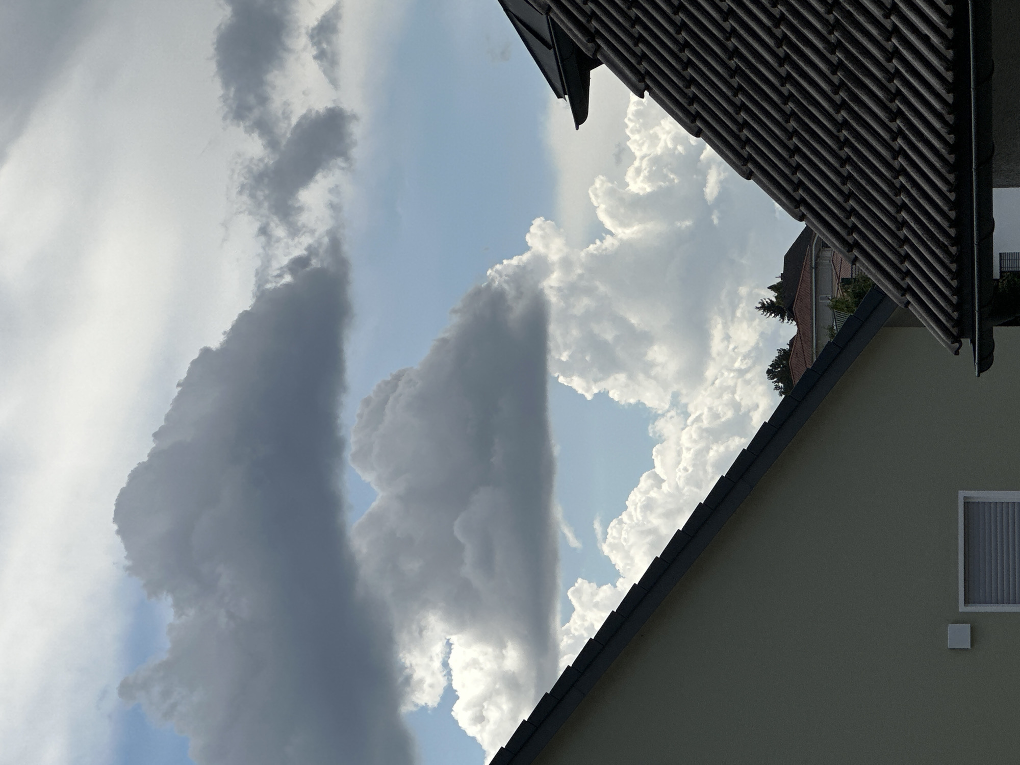 The image shows a view of the sky with dramatic clouds, partially obscured by the roofs of houses. In the foreground, the angled roofs of two houses are visible. The house on the left has a light-colored exterior wall and a dark-pitched roof with visible tiles. A rectangular window with closed blinds is present on this wall. The house on the right has a dark-tiled roof with a distinct eaves detail. In the background, more houses with red roofs can be seen, along with some trees. The sky is filled with a mix of dark, heavy clouds and bright, puffy cumulus clouds, suggesting a dynamic weather system. The scene is likely a residential area. Without further context, the specific location in Günzburg, Germany cannot be determined beyond it being a residential setting. There are no people or animals visible in the image, and no actions or interactions are depicted. The environment is characterized by domestic architecture and a cloudy sky.