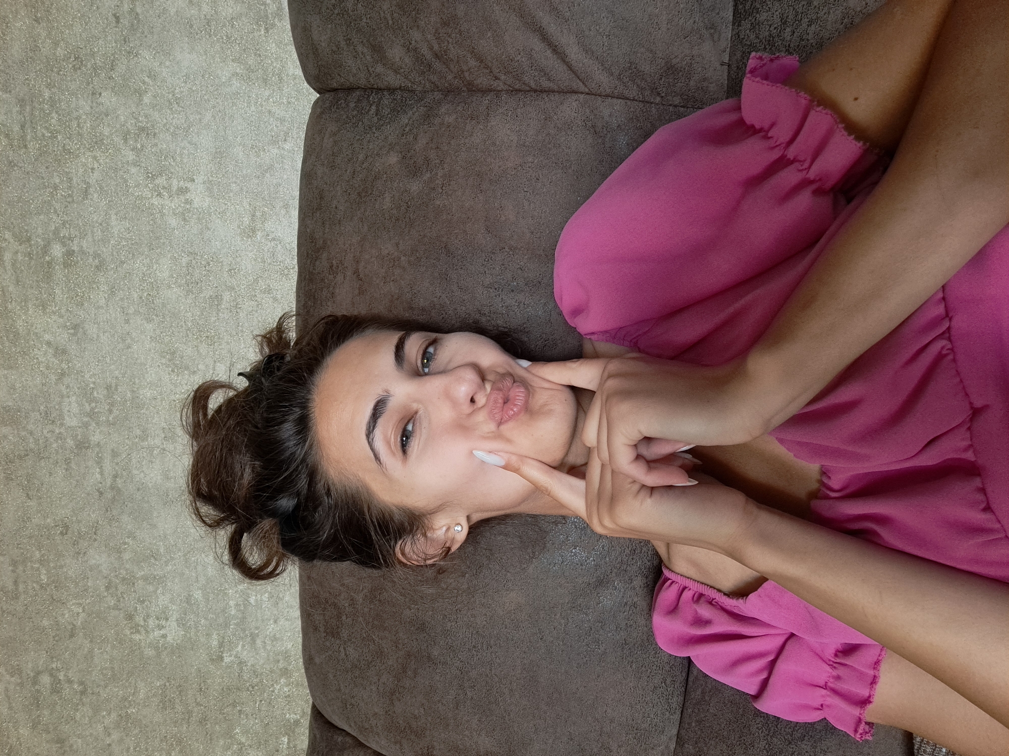 A young woman with brown hair pulled up in a messy bun and light-colored eyes is looking at the camera with puckered lips, her fingers touching her cheeks. She is wearing a bright pink top with ruffled sleeves and a square neckline. She is lying on a brown couch, with a textured wall behind her. The lighting is natural and appears to be from an overhead source.