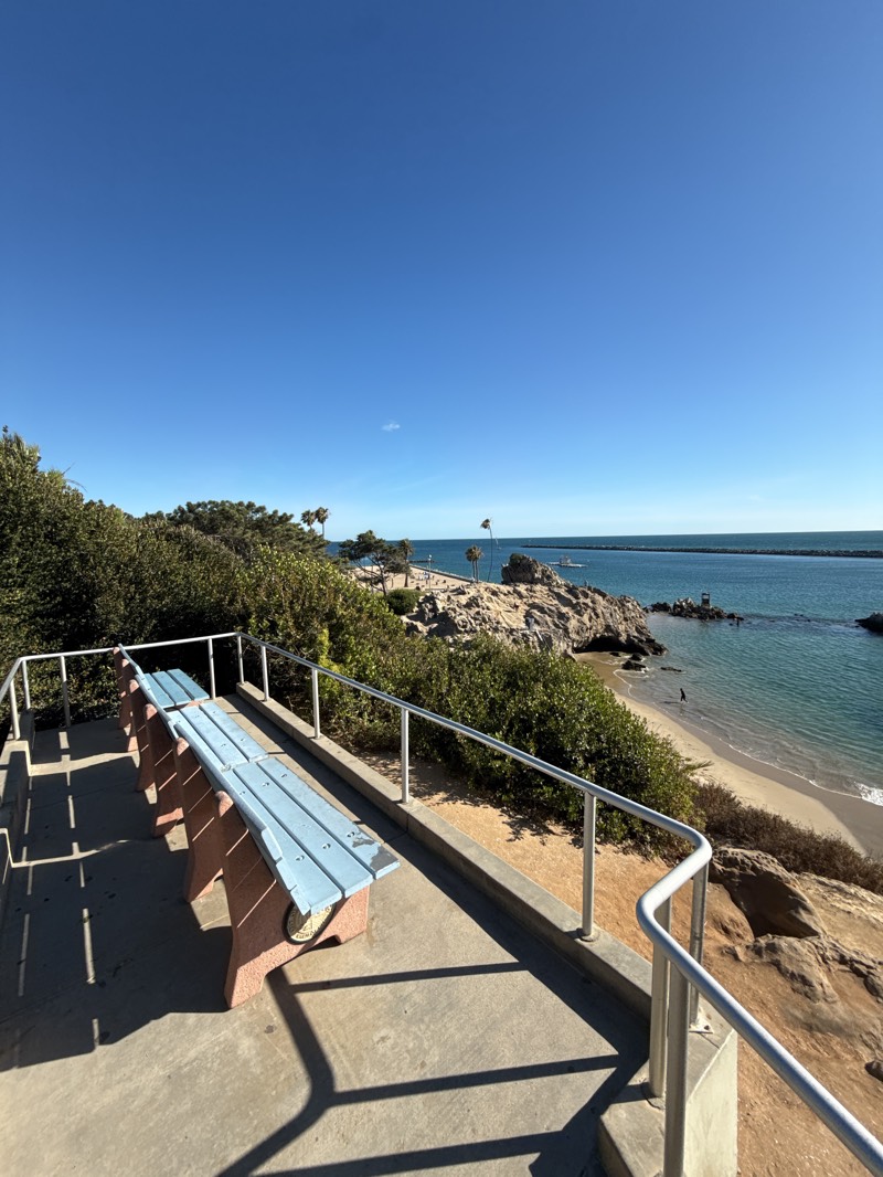 The image depicts a serene coastal scene in Newport Beach, United States, on a clear, sunny day. A weathered light-blue bench sits on a concrete platform enclosed by a silver railing. The platform overlooks a small sandy beach, turquoise water, and rocky cliffs covered with vegetation. In the distance, a jetty stretches out into the ocean. The sky is a deep, cloudless blue. There are no people clearly visible in the image.