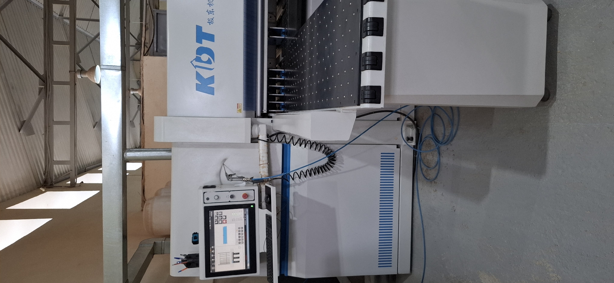 The image shows a KDT brand CNC machine in what appears to be a workshop or factory setting in Aktau, Kazakhstan. The machine is white with blue accents and has a digital interface with a keyboard and mouse. A spray nozzle is attached to the machine, and various cables and hoses are connected to it, running along the floor. The machine's work surface is visible, with a pattern of small dots. The background includes a corrugated metal roof, support beams, and ventilation pipes, suggesting an industrial environment. There are no people visible in the image. The floor is concrete and shows some dust or debris.