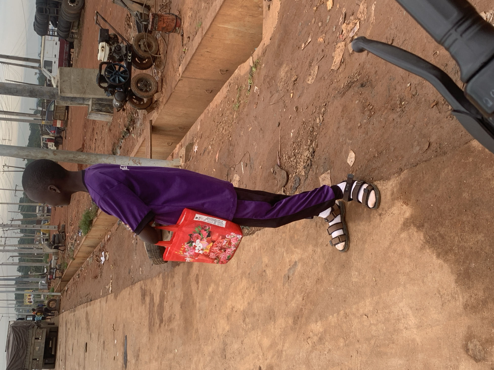 A young boy stands on a dirt road in Shagamu, Nigeria, holding a red bag with a floral pattern and a cartoon character. He is wearing a purple t-shirt with black and purple striped pants and brown sandals with white socks. In the background, a truck is parked on the left, and various vehicles, including a white van and a dark blue car, are visible. There are stacks of tires and a small engine with wheels. The environment appears to be an unpaved area with reddish-brown dirt, scattered debris, and a concrete drainage channel.
