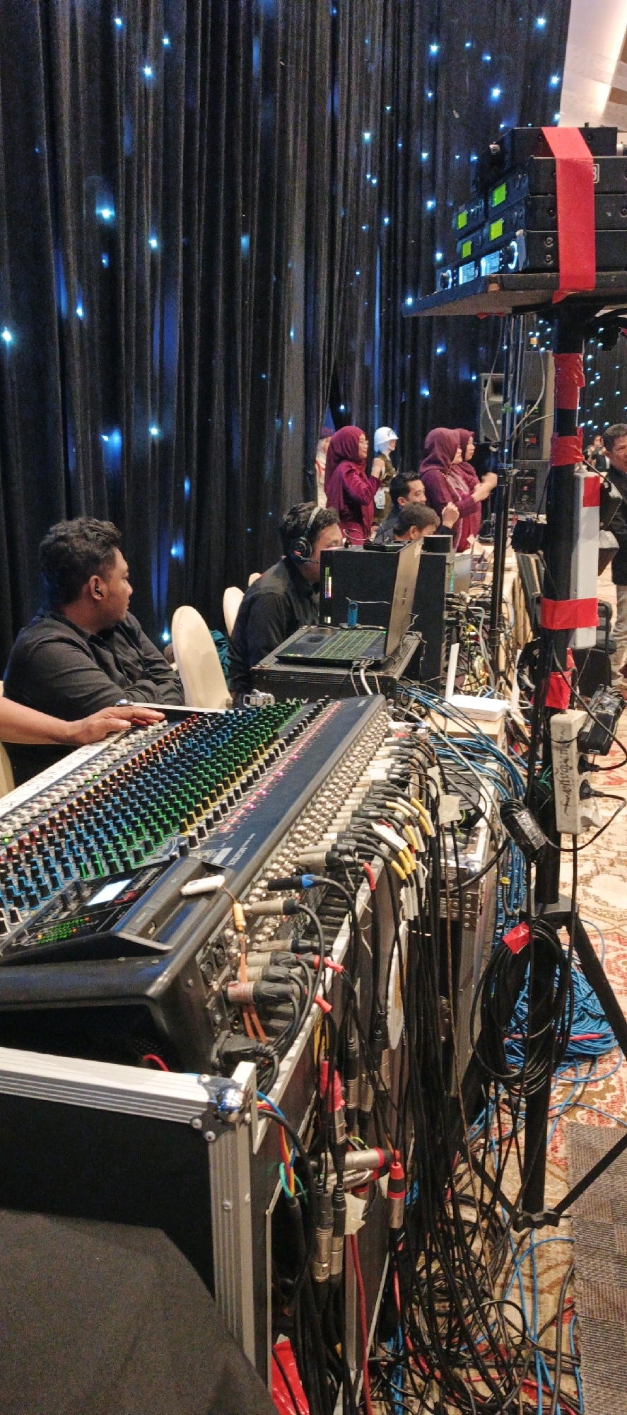 This is an outdoor, low-angle, full shot of a man in a black shirt operating a large audio mixing board in a dimly lit room, possibly a banquet hall. The room has a dark curtain backdrop with scattered blue lights, creating a starry effect. In the foreground, a large, multi-channel audio mixing board dominates the frame, with its colorful knobs and faders clearly visible. Cables of various colors snake across the floor and connect to the board and other equipment. To the left of the mixing board, another man in a black shirt is seated, with his hands on the board, seemingly engaged in the audio mixing process. Behind him, several individuals, including women wearing hijabs, are standing and appear to be observing or participating in an event. In the background, a rack of audio equipment, including wireless receivers, is mounted on a stand. The overall impression is that of a live event being managed by audio technicians. The image does not provide any explicit visual cues to pinpoint the exact location in Surabaya, Indonesia.