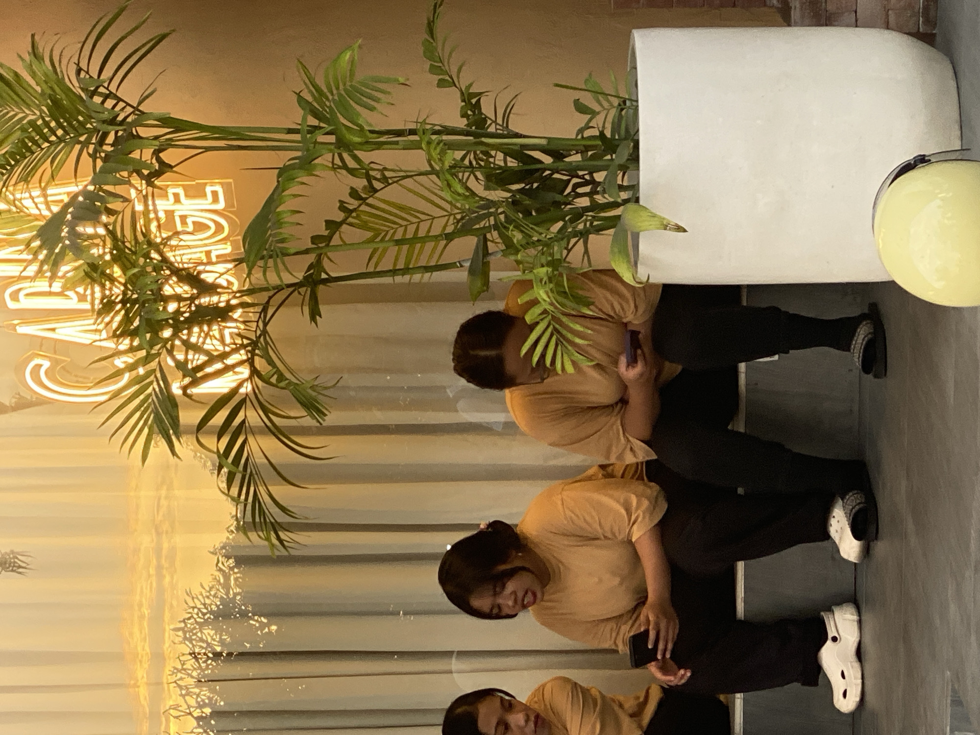 In Kuta Selatan, Indonesia, three individuals are seated on a stepped surface in front of a beige wall. To the left, a person wearing a tan shirt and black pants, with dark hair pulled back, holds a black smartphone. Next to them, another person in a tan shirt and black pants, with dark hair styled in a bun, also holds a black smartphone. To the far right, a third individual, partially obscured and wearing a tan shirt and black pants, is also looking at a smartphone. They are all wearing casual footwear; one has white Crocs, another has black slides with white accents, and the third person's footwear is less visible. In the foreground, a large white planter contains a leafy green plant. A yellow helmet rests on the floor in front of the planter. Behind the individuals, sheer curtains are illuminated by a warm light, and neon lights forming a word, possibly "MASSAGE," are visible in the upper background. The overall scene appears to be an indoor waiting area or lounge.