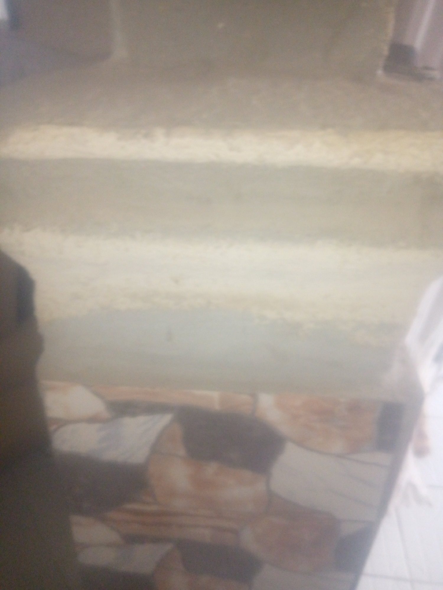 The image is a blurry, medium-angle shot of what appears to be a staircase. The staircase has multiple steps, with each step having a lighter colored horizontal band. The surface of the steps appears textured and somewhat dusty. In the foreground, there is a patterned floor, possibly tiled, with irregular shapes in shades of brown and white. To the left of the frame, a dark, indistinct shape, possibly a person or an object, is visible. To the right, a blurry white object, possibly clothing or fabric, is present. The overall lighting is dim and diffused, contributing to the low clarity of the image. The location is Jauro Yinu, Nigeria.
