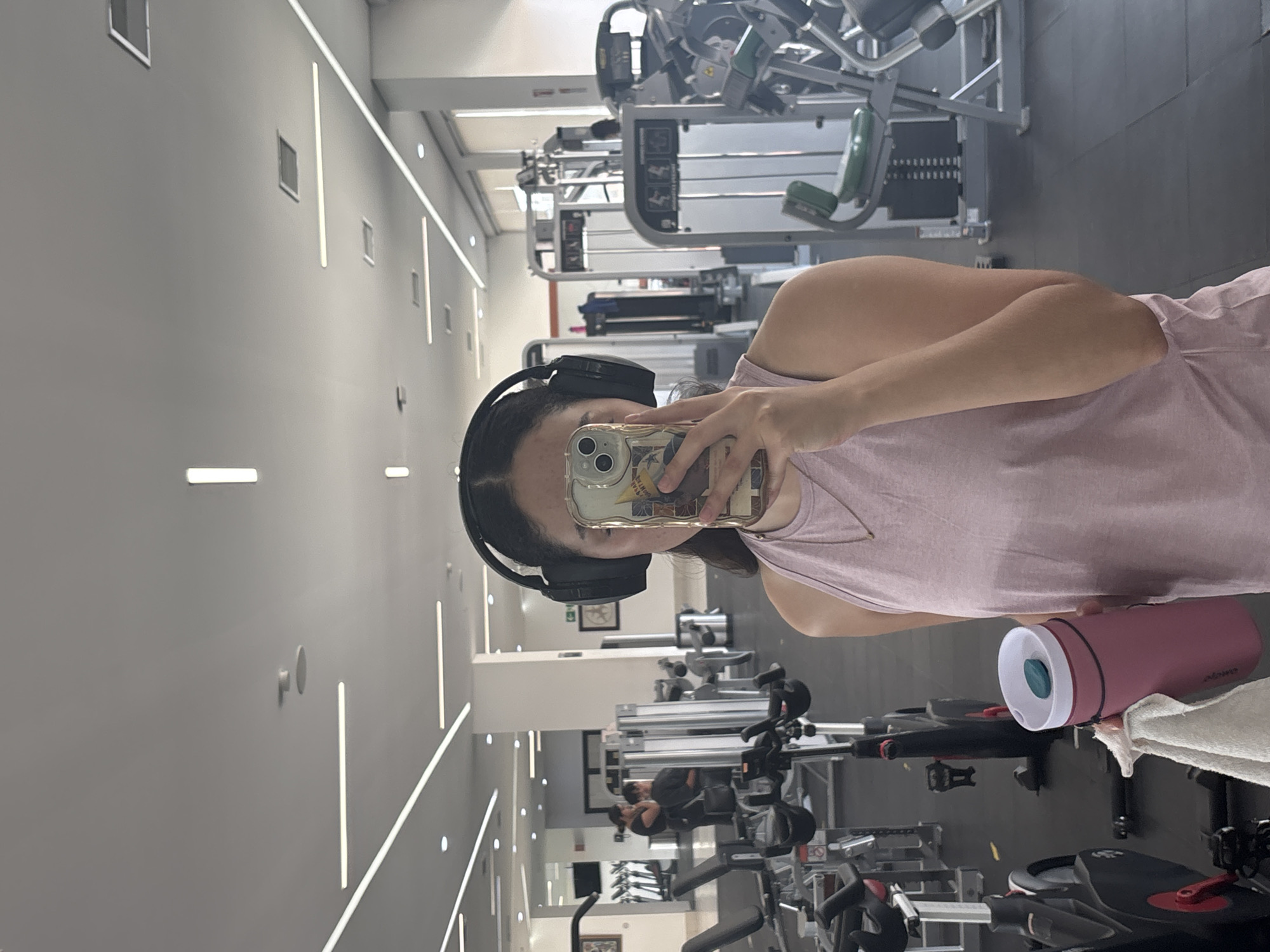 A person in a gym setting holds a phone up to take a selfie, with a water bottle and towel visible in the foreground. The gym contains various exercise machines and equipment. The gym's walls are light gray, and the interior lighting is bright. Based on available information, the location is San Andrés Cholula, Mexico.