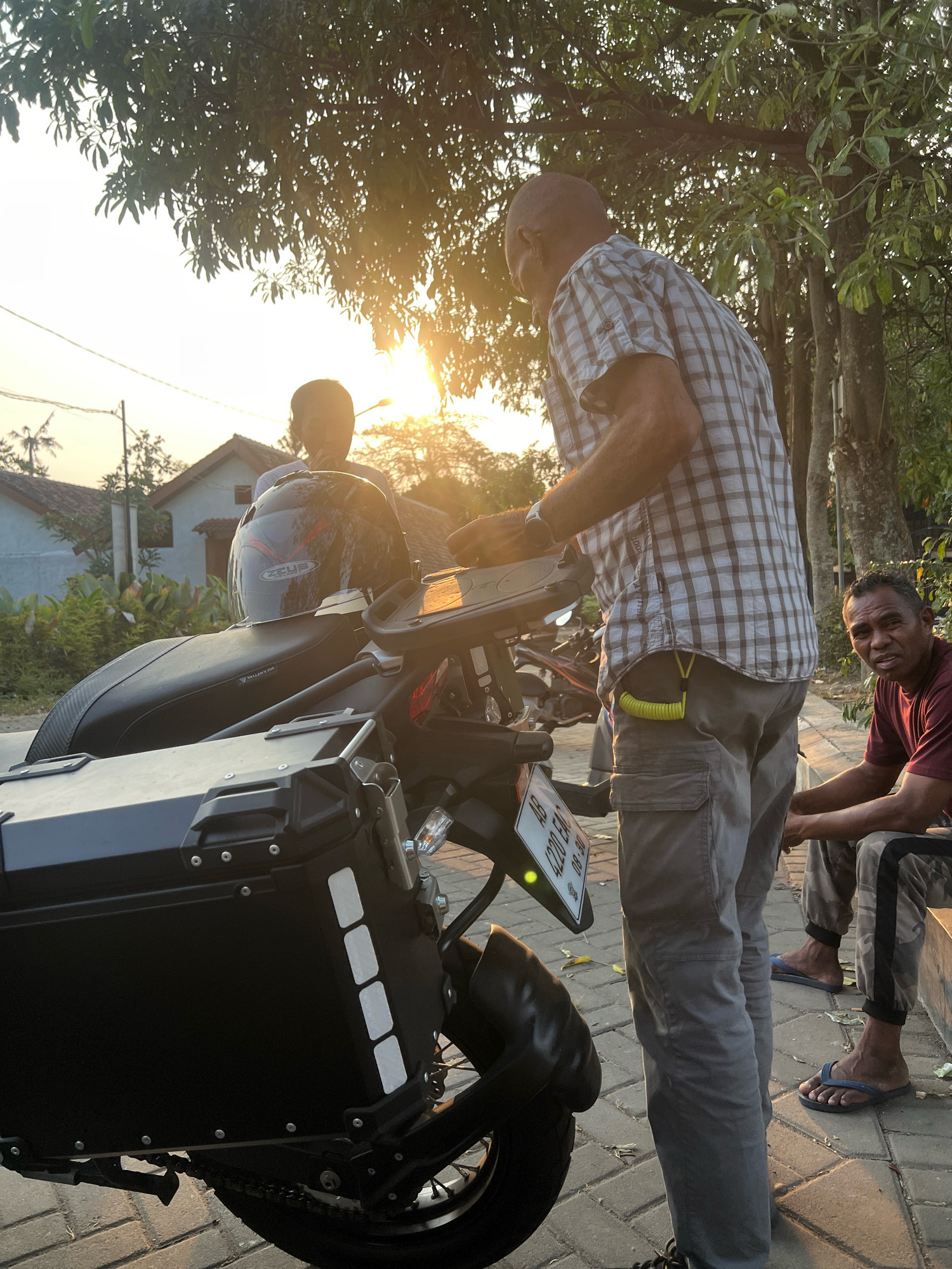 In a scene set in Ngaglik, Indonesia, two men are pictured near a motorcycle during what appears to be golden hour. The man on the left, partially obscured by the motorcycle, wears a helmet. The man in the center, who is taller and standing, is dressed in a checkered shirt and gray cargo pants. He is interacting with the motorcycle's dashboard. A second man, seated to the right, wears a maroon t-shirt and camouflage pants, looking towards the camera. The motorcycle is equipped with large black panniers on the rear. The background features residential buildings and lush trees, illuminated by the warm glow of the setting sun. The ground is paved with bricks.