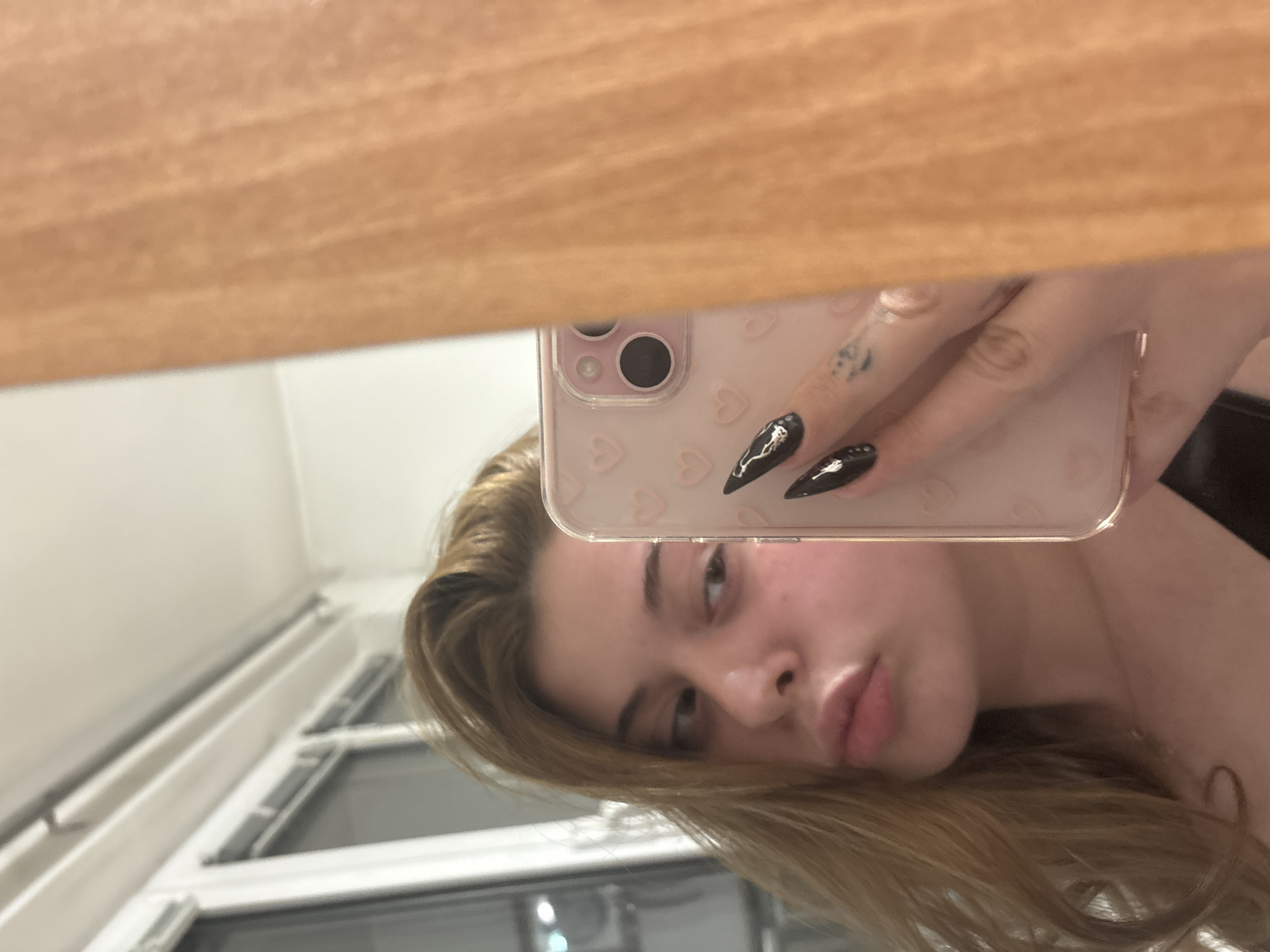 The image shows a woman holding up a pink iPhone with a heart-patterned case, taking a selfie in a mirror. The woman has light brown hair, visible from her shoulders up. Her black nails with white designs are visible in the foreground. The reflection shows a room with a window, suggesting an indoor setting. The top of the mirror frame is visible at the top of the image. The location in Troyan, Bulgaria cannot be determined from the image.