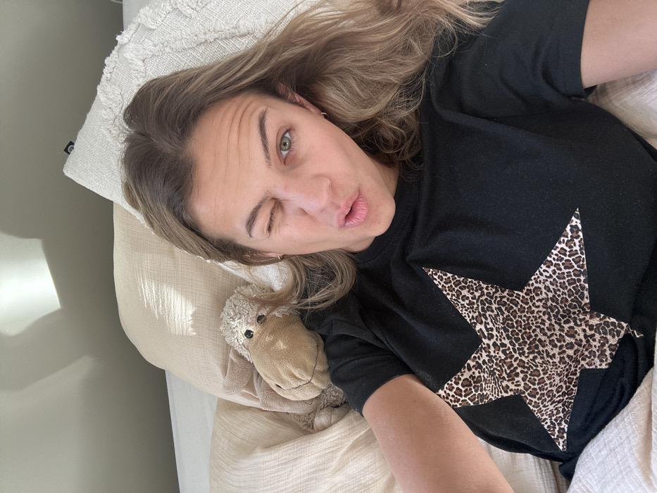 A female subject is depicted lying in bed in a room within Veauchette, France. She has light brown hair, is wearing a black t-shirt with a leopard-print star design on the chest, and small hoop earrings. Her right eye is closed in a wink, and her lips are puckered. A light-colored stuffed animal, resembling a sheep, lies next to her left shoulder on a light beige pillow. Additional pillows, one with a textured pattern, and a light-colored duvet are visible. A light gray wall forms the background, exhibiting a bright rectangular reflection consistent with natural light from an unseen window.
