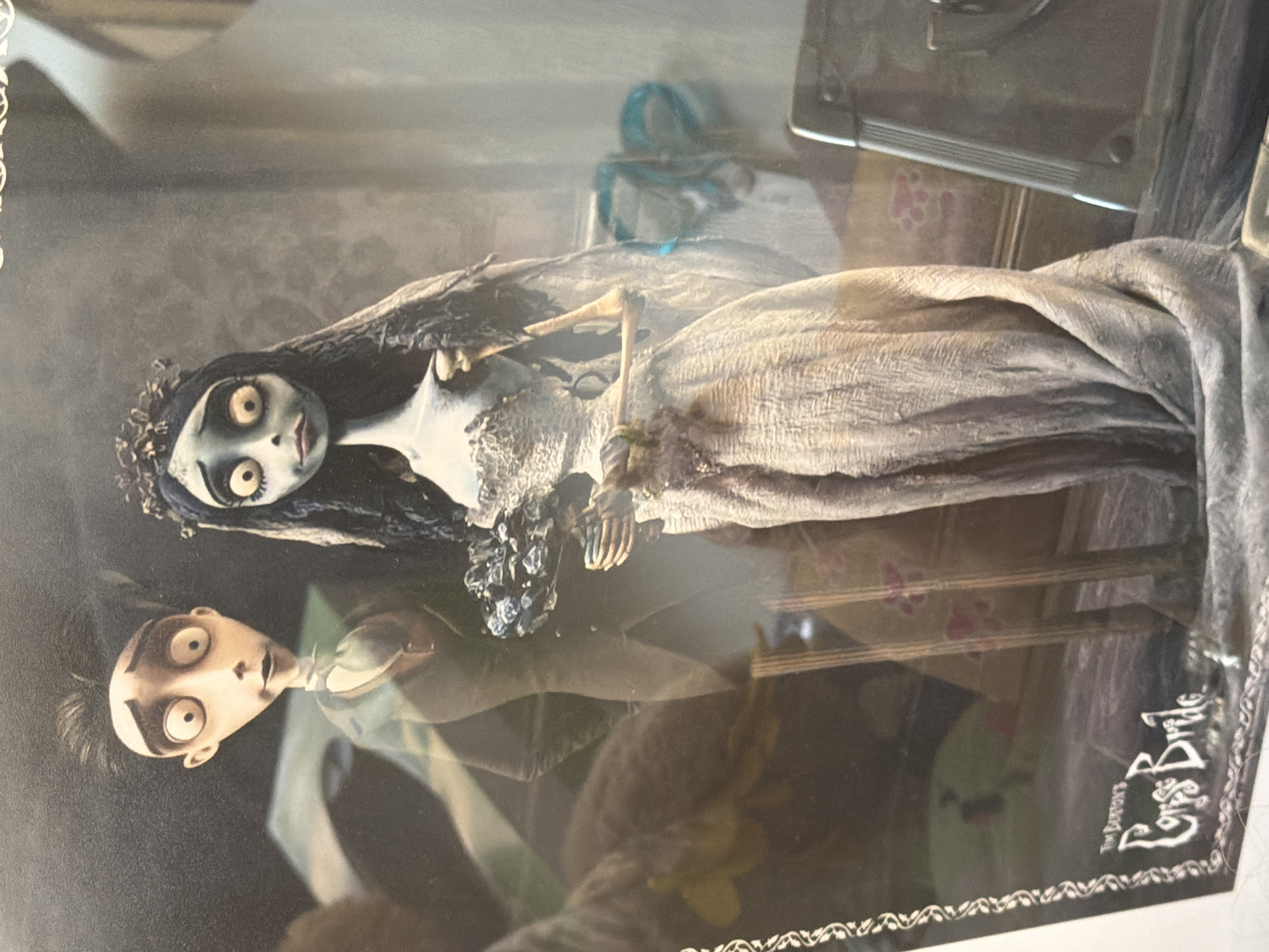 The image displays two stylized figures, reminiscent of characters from stop-motion animation, positioned in a somewhat eerie, gothic setting. The figure on the left appears to be a man with wide, startled eyes, dark spiky hair, and wearing a dark suit with a white cravat. The figure on the right is a woman with pale blue skin, large, dark-rimmed eyes, black hair cascading down, and wearing a tattered, white wedding dress adorned with delicate lace. She has a flower bouquet clutched in her hands. The background is dark and appears to be a room with textured wallpaper and some indistinct objects. The overall impression is that of a display or a promotional image for a film or product, likely related to "Tim Burton's Corpse Bride," as evidenced by the partially visible text at the bottom left of the image. There is no discernible location in Alicante, Spain, or any other specific geographic location visible in the image.