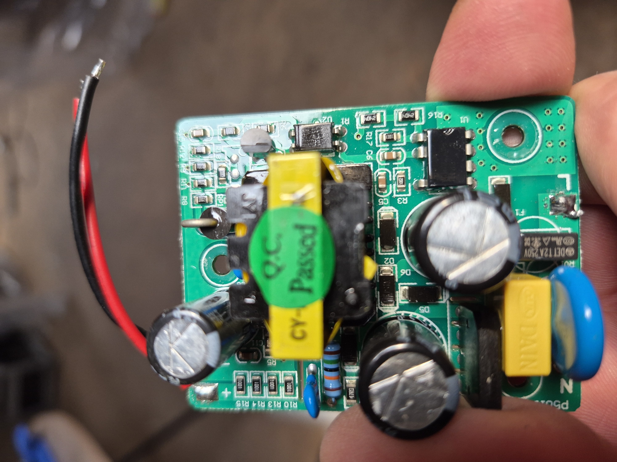The image shows a close-up, slightly blurry view of a green printed circuit board (PCB) with various electronic components. The PCB is held in the palm of a person's hand. Prominent components include several electrolytic capacitors (one with a silver top and a cross-shaped indentation, two larger ones with black casings and silver tops), a yellow film capacitor with "CY- Passed" printed on it in green, a black chip with multiple pins, resistors, and surface-mount components. Red and black wires are connected to the top of the PCB. The background is out of focus and appears to be an indoor setting with a blurred wooden surface. There is no visual information in the image that relates to the city of Masów, Poland.