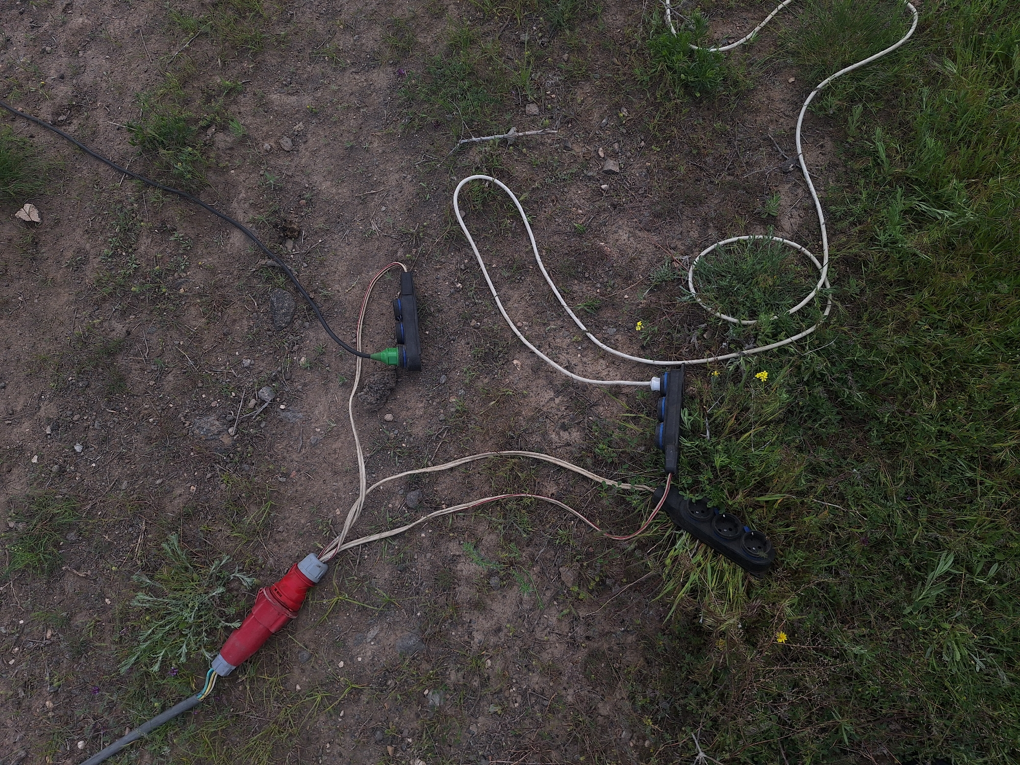 The scene depicts an unidentifiable location with ground covered by a mix of dirt and grass. Several electrical components are scattered across the area. A large, red industrial plug is connected to a gray cable, extending towards the lower-left corner. Two black power strips are visible, each with three sockets. One strip is connected to a green plug, while the other is linked to a white cable that loops around in a coil. No people are present in the frame. The location in Tlik, Armenia is not identifiable from the given image.