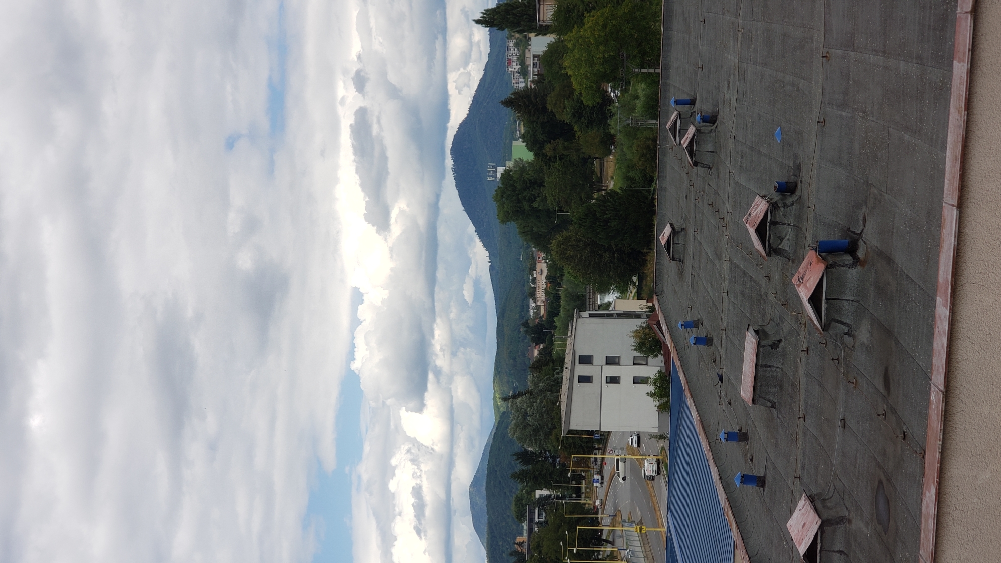 The image shows a rooftop view of a city with rolling hills and a large mountain in the background, likely in or near Prešov, Slovakia. The foreground consists of a flat, dark grey rooftop covered in what appears to be asphalt shingles. Several ventilation shafts with blue cylindrical bases and reddish-brown, weathered conical caps are scattered across the roof. There are also several smaller, triangular structures with similar reddish-brown caps, possibly skylights or access hatches. The middle ground features a modern, white, rectangular building with multiple windows. To the left of this building, a road with traffic can be seen, including several cars and a white van. Trees and more buildings are visible in the distance, nestled amongst the greenery. The background is dominated by lush green hills and a prominent, dark, conical mountain with some structures on its peak. The sky is overcast with a mix of grey and white clouds, with patches of blue sky visible. The overall scene depicts an urban environment bordering a natural landscape.