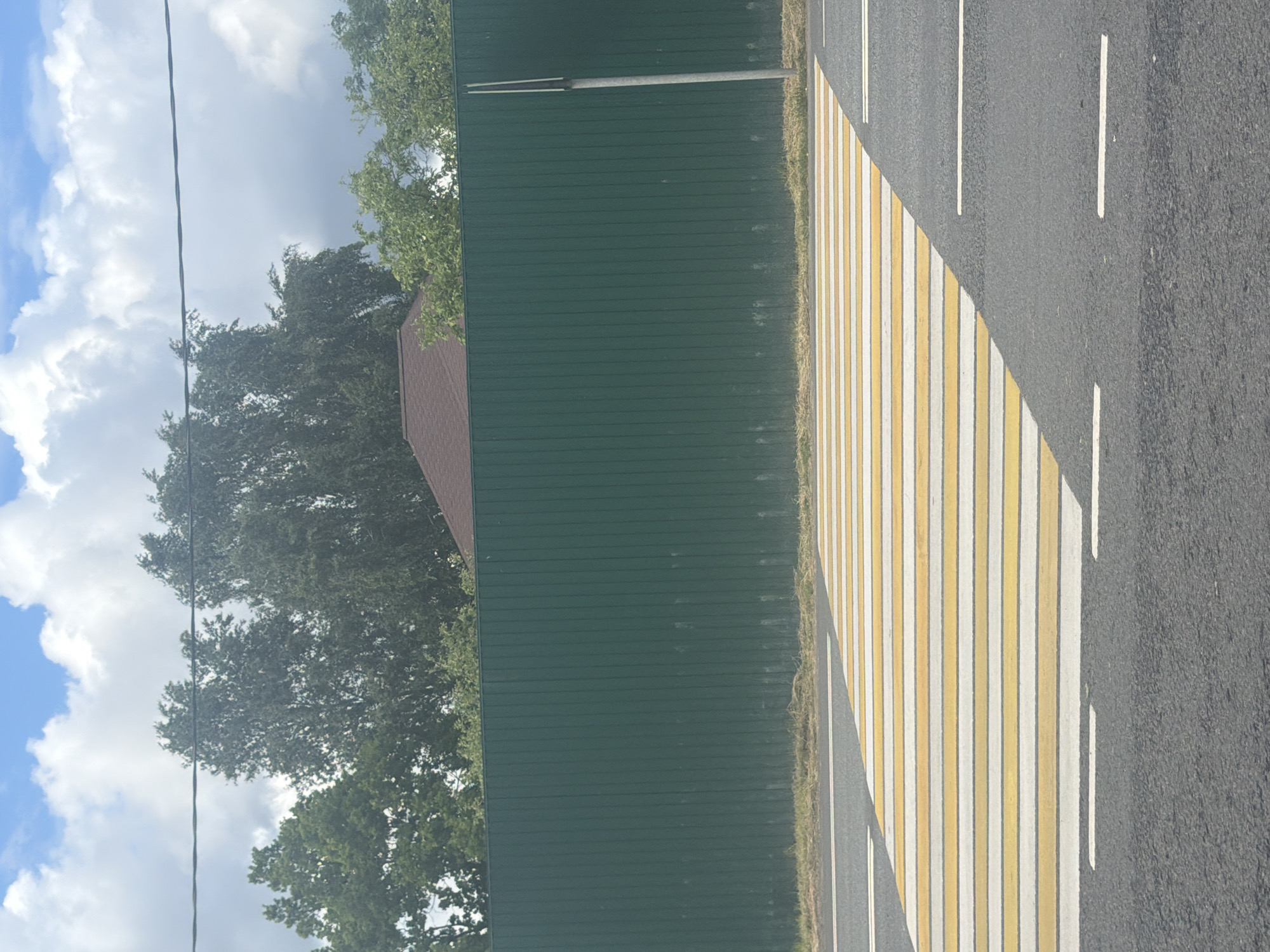 The image depicts a street in Borki, Russia. The view is from the roadside, looking towards a section of asphalt pavement marked with white and yellow pedestrian crossing stripes. Adjacent to the crossing is a green metal fence with horizontal slats, partially obscured by trees. A section of a house roof can be seen behind the fence. Overhead, the sky is visible with white clouds. The road also has intermittent white lane divider stripes.