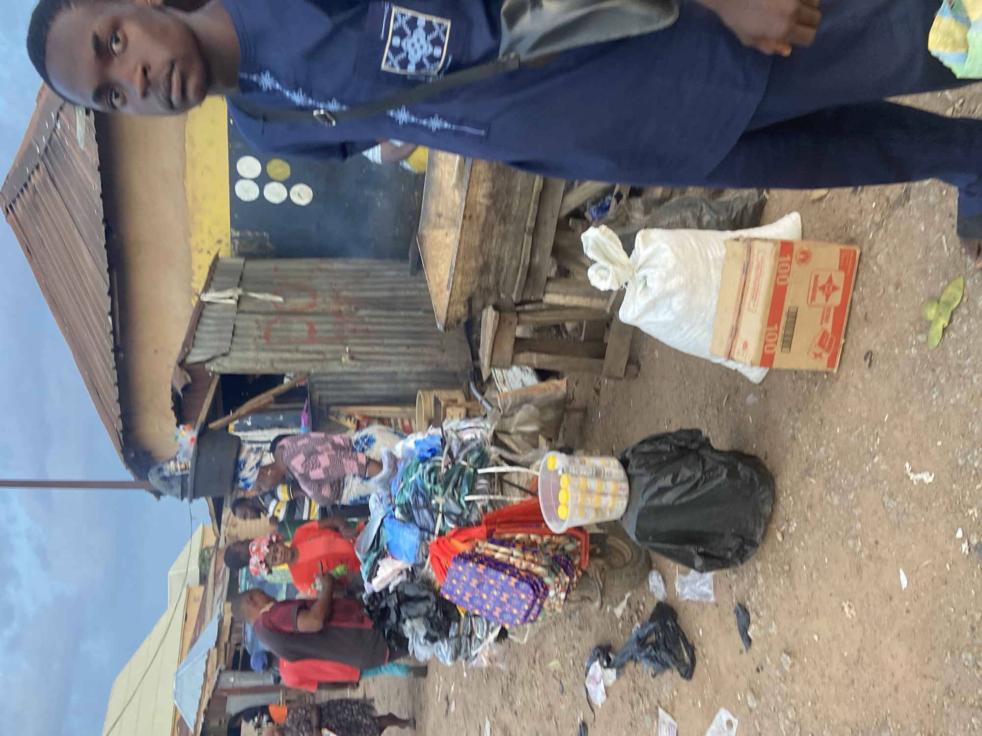 In Bukuru, Nigeria, a street market scene unfolds. A young man in a blue garment stands in the foreground, his gaze directed forward. Behind him, a cart is laden with various items, including bags and what appear to be packaged goods. Several other individuals are present, some engaged in commerce or interacting with the goods on display. The environment suggests a bustling marketplace with makeshift stalls and a dusty, outdoor setting. The sky above is overcast.