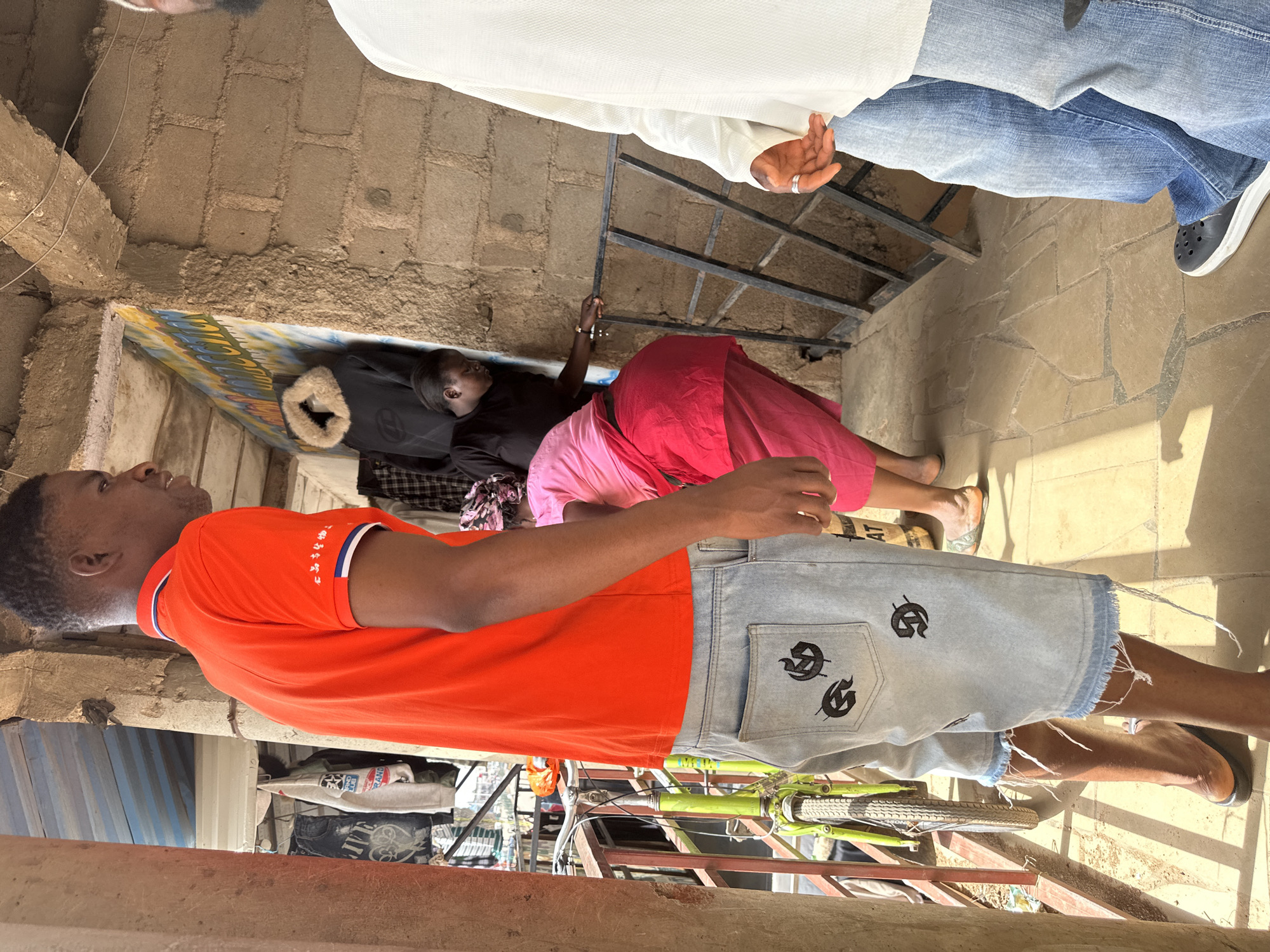 In Bukuru, Nigeria, a man in an orange polo shirt and distressed denim shorts stands on a paved walkway. To his right, another man in a white shirt and blue jeans is partially visible. Behind the first man, a woman in a black shirt and pink skirt is seen entering a doorway with a fur-lined coat hanging inside. Another woman, wearing a pink headscarf and pink top, is positioned in front of the doorway. A green bicycle is propped against a railing to the left. The environment appears to be an outdoor market or residential area, with exposed brick walls, corrugated metal roofing, and various items hanging on display in the background. The sunlight casts shadows on the ground.