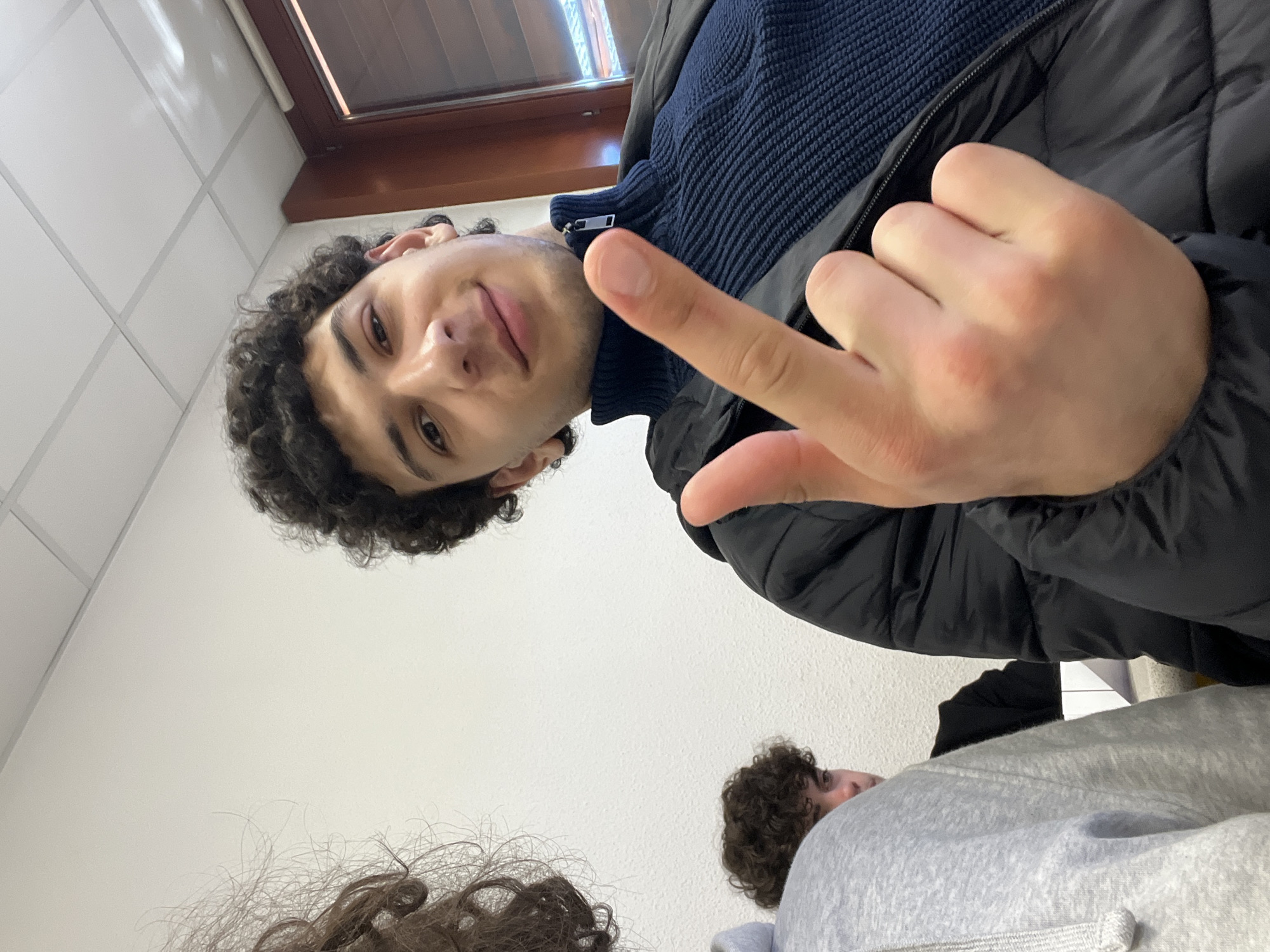 A selfie-style image of a young man with dark curly hair, wearing a navy blue turtleneck sweater and a black puffer jacket. He is holding up his middle finger. Two other individuals with curly hair are partially visible at the bottom of the frame. The background suggests an indoor setting, possibly a classroom, with white tiled walls and a window in the upper portion of the image. The location in Schiltigheim, France, is not discernible from the visual data.
