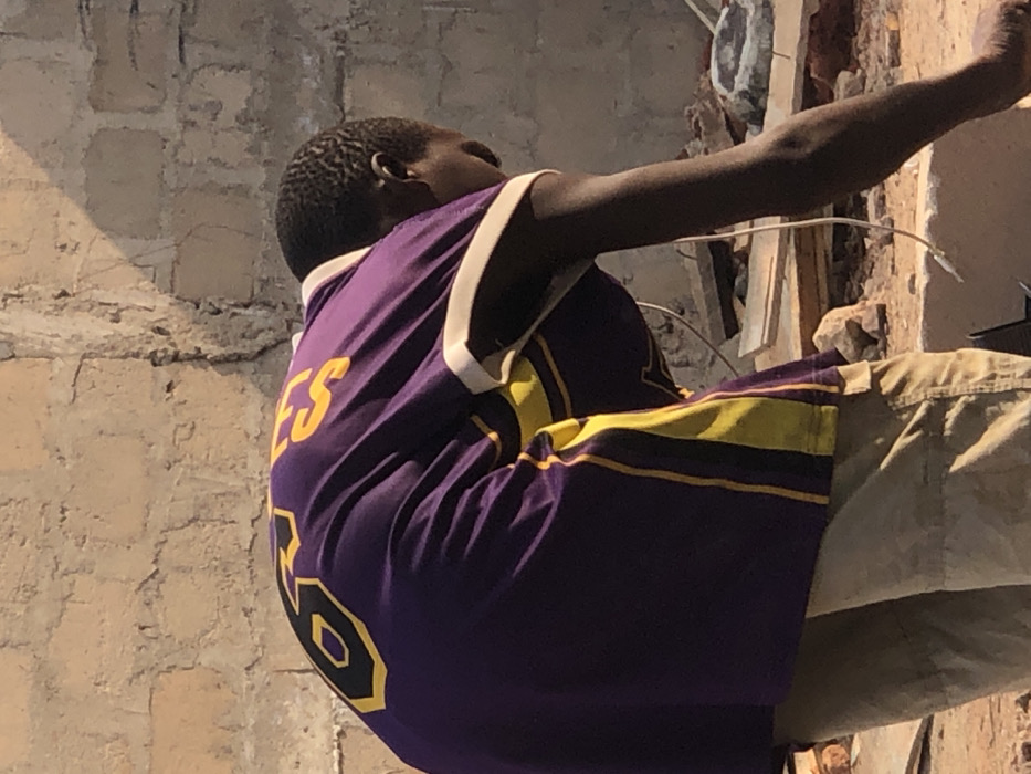 A male individual with dark skin and short dark hair is depicted bending forward and looking downwards, engaged in an unspecified activity. He is wearing a purple short-sleeved shirt with yellow and black numbers "8" and letters "ES" visible on the back, along with yellow vertical stripes on the sides and white trim on the collar and sleeve ends. A white cable, consistent with a headphone or charging cord, hangs from his right side near the ear area. He wears tan-colored pants or shorts. The scene is outdoors, set against a background of a light-colored, weathered concrete block wall showing visible mortar lines and some cracking. The ground beneath him is uneven, dusty, and strewn with construction debris, including rocks, pieces of wood, and fragmented materials. The lighting suggests strong sunlight from an overhead source, casting shadows on the wall and the ground. This scene takes place in Enugwu-Ukwu, Nigeria.