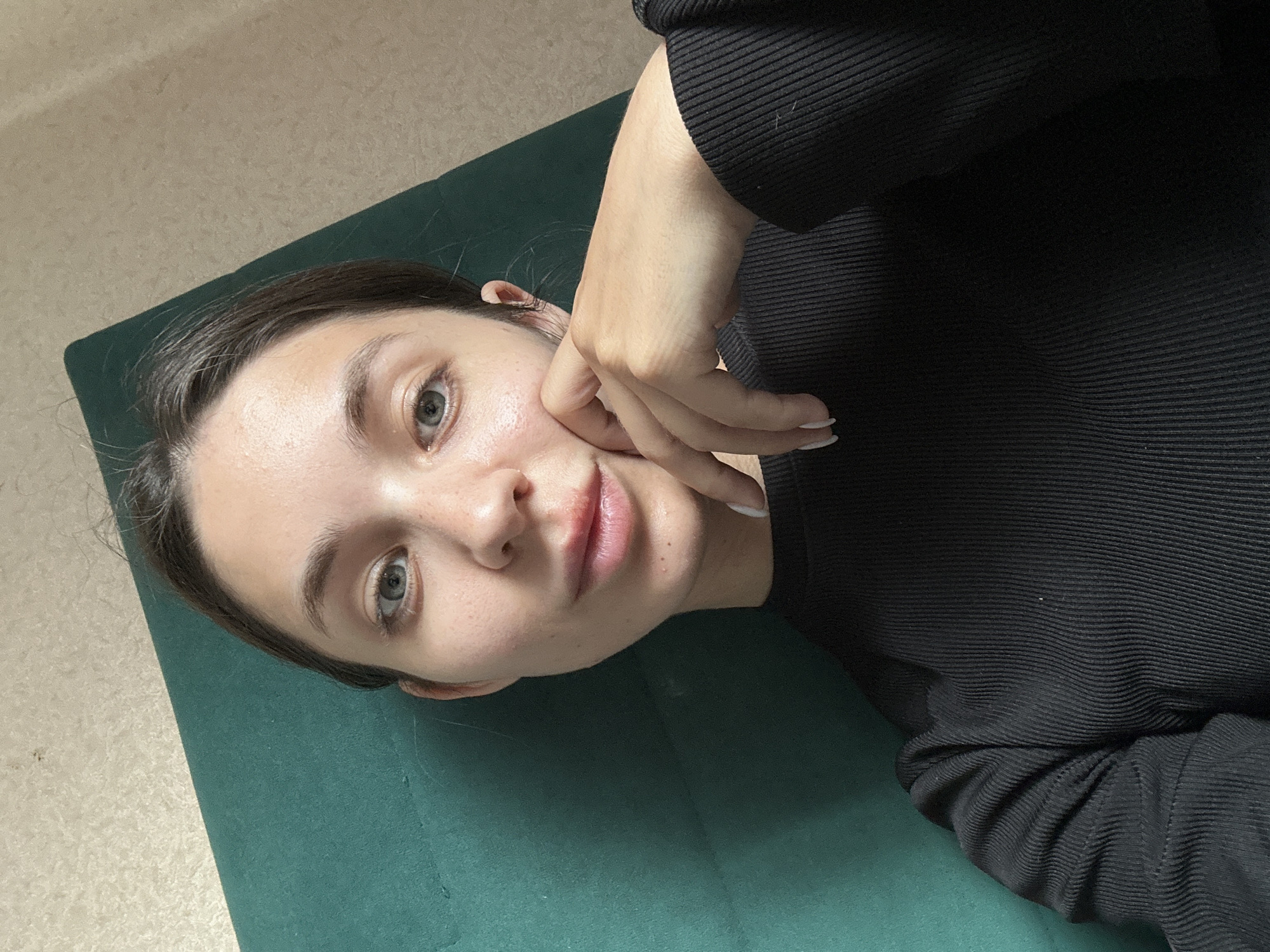 This media file features a close-up, overhead shot of a young woman lying down. She is wearing a black, ribbed long-sleeved shirt. Her face is turned towards the camera, and her left hand is gently touching her cheek. Her eyes are open and looking forward, and her lips are slightly parted. The woman has fair skin with some light freckles, and her eyebrows are dark. Her hair is dark brown and pulled back. She is lying on a dark green mat, which is situated on a light-colored, textured floor. The lighting suggests it might be daytime, with natural light illuminating her face. The overall mood appears relaxed and contemplative. There is no visible text in the image. The location context provided is Sverdlovskiy, Russia.