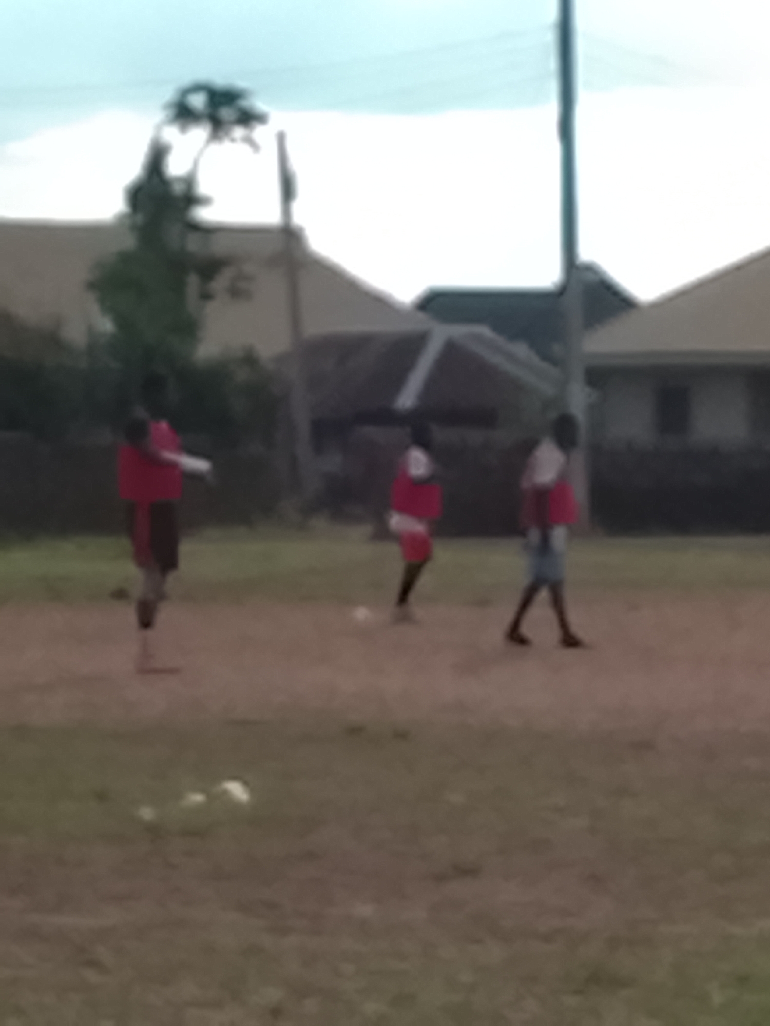 Three individuals, appearing to be young males, are present on a dirt field, possibly a football pitch, in Rafin Bauna, Nigeria. They are wearing red tops and dark shorts or pants. One individual on the left is in the process of kicking a ball, while the other two are standing, observing or awaiting the play. The environment includes some vegetation and buildings in the background. The lighting suggests it is daytime, possibly late afternoon or early morning.
