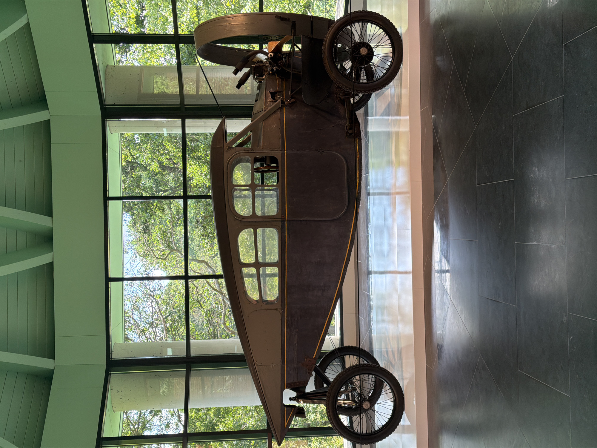 The image depicts a vintage, dark-colored automobile with a distinctive aerodynamic design, possibly a concept car or an early aircraft-inspired vehicle. It is displayed indoors, with large windows providing a view of lush green trees and dappled sunlight, suggesting a natural or museum setting. The car has two prominent spoked wheels at the front and a single large wheel at the rear, enclosed by a curved cowling. The interior appears to have seating for at least two occupants, with visible steering mechanisms. The vehicle is positioned on a white platform, which is set against a dark, reflective floor. The overall impression is one of a meticulously preserved artifact. This scene is located at the Louwman Museum in The Hague, Netherlands, a renowned institution dedicated to automotive history.