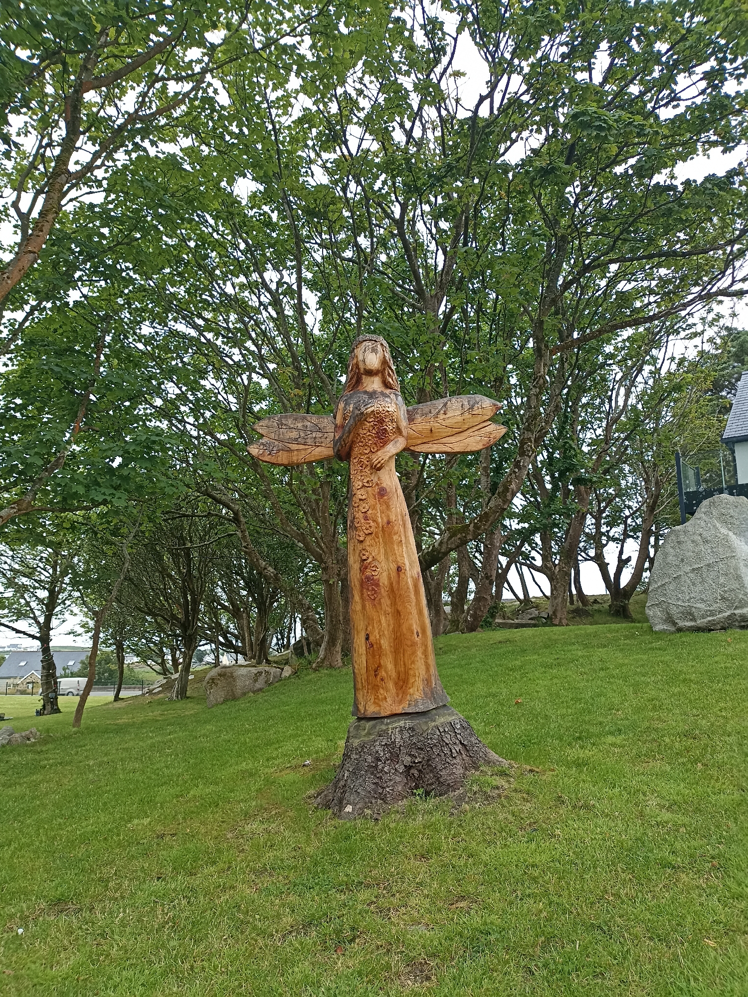 This is a medium-shot, eye-level, outdoor photograph of a wooden sculpture of an angel or winged figure. The sculpture is carved from a tree trunk and is positioned on a grassy slope, with a backdrop of lush green trees. The angel has carved wings extending from its back, and its body is adorned with floral patterns. It stands on a rough, naturalistic base, the remaining stump of the tree.
In the background, there are many trees with dense green foliage, suggesting a temperate climate and possibly a late spring or summer season. The grass in the foreground is short and well-maintained, indicating a park, garden, or public space. To the far left, a white building with a dark roof can be seen, and a white van is parked nearby, suggesting a residential or commercial area bordering the park. To the far right, a large grey boulder sits on the grass. The sky is overcast, implying a cloudy day, typical of the weather in Ireland. There is no discernible text in the image. The scene appears tranquil and serene, with the naturalistic sculpture harmonizing with the surrounding environment. The location is identified as Doirí Beaga, Ireland.