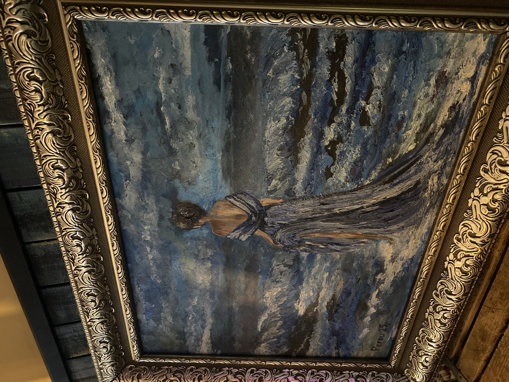 The image shows a framed oil painting of a woman in a blue gown standing by the ocean. The painting is in landscape orientation and features a dramatic sky and turbulent waves. The woman is facing away from the viewer, with her back to the ocean. Her gown is long and flowing, and appears to be made of a light, sheer fabric. The painting is signed "Pereli 25" in the lower left corner. The frame is ornate and gold-colored. The painting is hung on a dark blue, wooden paneled wall. The foreground shows a wooden surface, possibly a table or shelf, with some indistinct objects on it. There is no visible indication of the location being Poltava, Ukraine.
