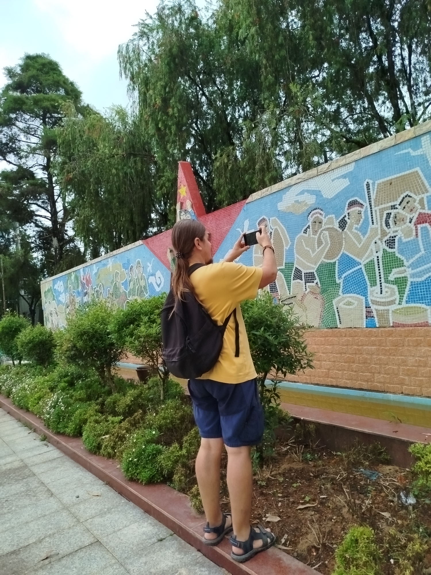 The image depicts an outdoor scene in what appears to be a park or public area in Cam Ly, Đà Lạt, Vietnam. A person, seen from behind, is the primary subject, standing on a paved pathway and actively photographing a colorful mosaic mural with a black smartphone held horizontally. The person has long brown hair tied in a ponytail, wears a yellow t-shirt, blue shorts, and dark sandals with "QUECHUA" visible on the strap of the right sandal. A black backpack is worn on their back.
The mosaic mural, composed of numerous small, colored tiles, stretches across a long wall. It illustrates stylized human figures engaged in various activities, utilizing a palette of blues, greens, browns, whites, and reds. The wall itself features a light blue top border and a brownish-orange brick-patterned base, with a shallow channel or pool of yellowish water running along its bottom. To the right of the mural, a distinct red, star-shaped structure is partially visible.
In front of the mosaic wall, a row of well-maintained green bushes and smaller white-flowering plants lines a landscaped bed. Beyond the wall, tall trees with dense green foliage, some exhibiting drooping branches, dominate the background. The sky is overcast or lightly cloudy, indicating a daytime setting with diffused light. The scene suggests a cultural or recreational space.
