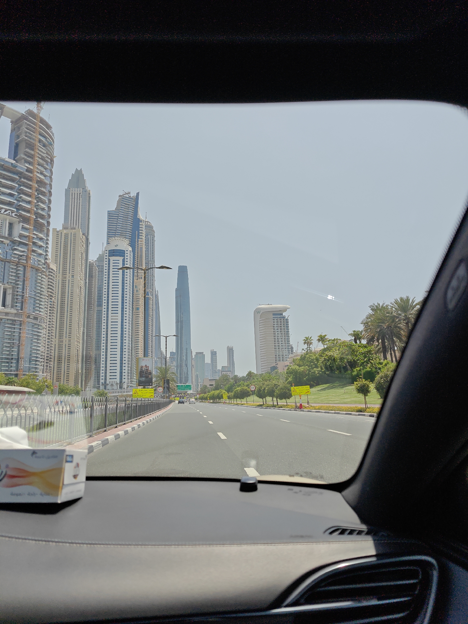 The image, taken from inside a vehicle, shows a view of Dubai Marina. Tall skyscrapers dominate the skyline on the left, with several under construction. A multi-lane road stretches ahead, bordered by a metal fence and a green park area with palm trees on the right. Traffic lights and street signs are visible along the road. The sky is clear and bright, suggesting a sunny day. In the foreground, a box of tissues rests on the car's dashboard. The overall scene depicts a modern urban landscape in Dubai.