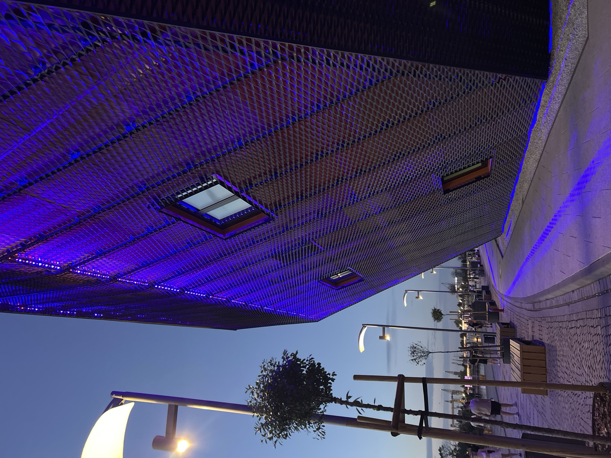An outdoor scene captures a modern, angled building with a perforated metal facade illuminated by purple LED lights at dusk. The building's design features geometric patterns and several small, rectangular windows. In the foreground, a paved walkway is lined with wooden benches and trees. The walkway is illuminated by blue lights integrated into the ground along its edge, creating a vibrant glow. To the left, a tall lamp post with a large, white, curved shade stands near a young tree supported by wooden stakes. In the background, a body of water with boats and cranes is visible, suggesting a harbor or waterfront area. Several people are walking along the pathway, some observing the building and others engaged in conversation. The sky is a deep twilight blue, indicating either early evening or late dusk. This scene is located at the Wały Chrobrego (Chrobry Embankment) in Szczecin, Poland, a prominent waterfront promenade known for its architecture and public spaces.