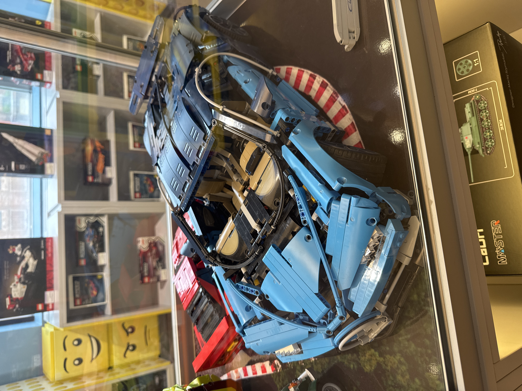The image displays a collection of LEGO models in what appears to be a retail display case. The primary subject is a large, partially assembled blue LEGO supercar, resembling a Bugatti Chiron, with its intricate mechanical details exposed. Next to it, a red LEGO sports car is visible. In the background, shelves contain various other LEGO set boxes, including a white and red Formula 1 car, a rocket, and an orange sports car. The display case is illuminated, reflecting on the surfaces of the LEGO models and the glass. The lower portion of the image shows a box with a detailed illustration of a green military tank, labeled "1:1" and "26.6CM". The context suggests this is a toy store or a hobby shop. There is no information in the image to indicate a specific location within Yekaterinburg, Russia.