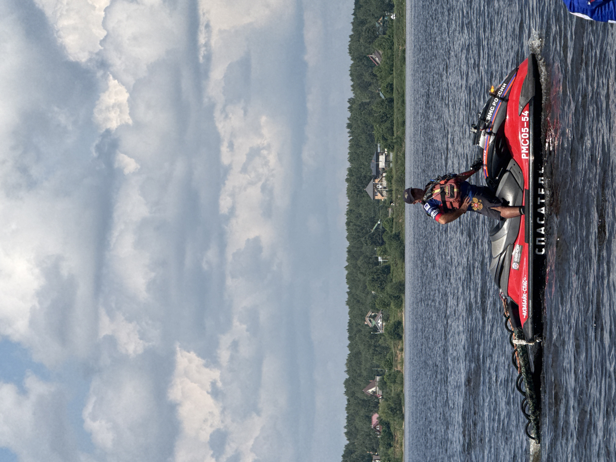 A man in a life vest stands on a red jet ski in the water. The jet ski has Russian writing on it. In the background is a tree line and some houses. The sky is cloudy.