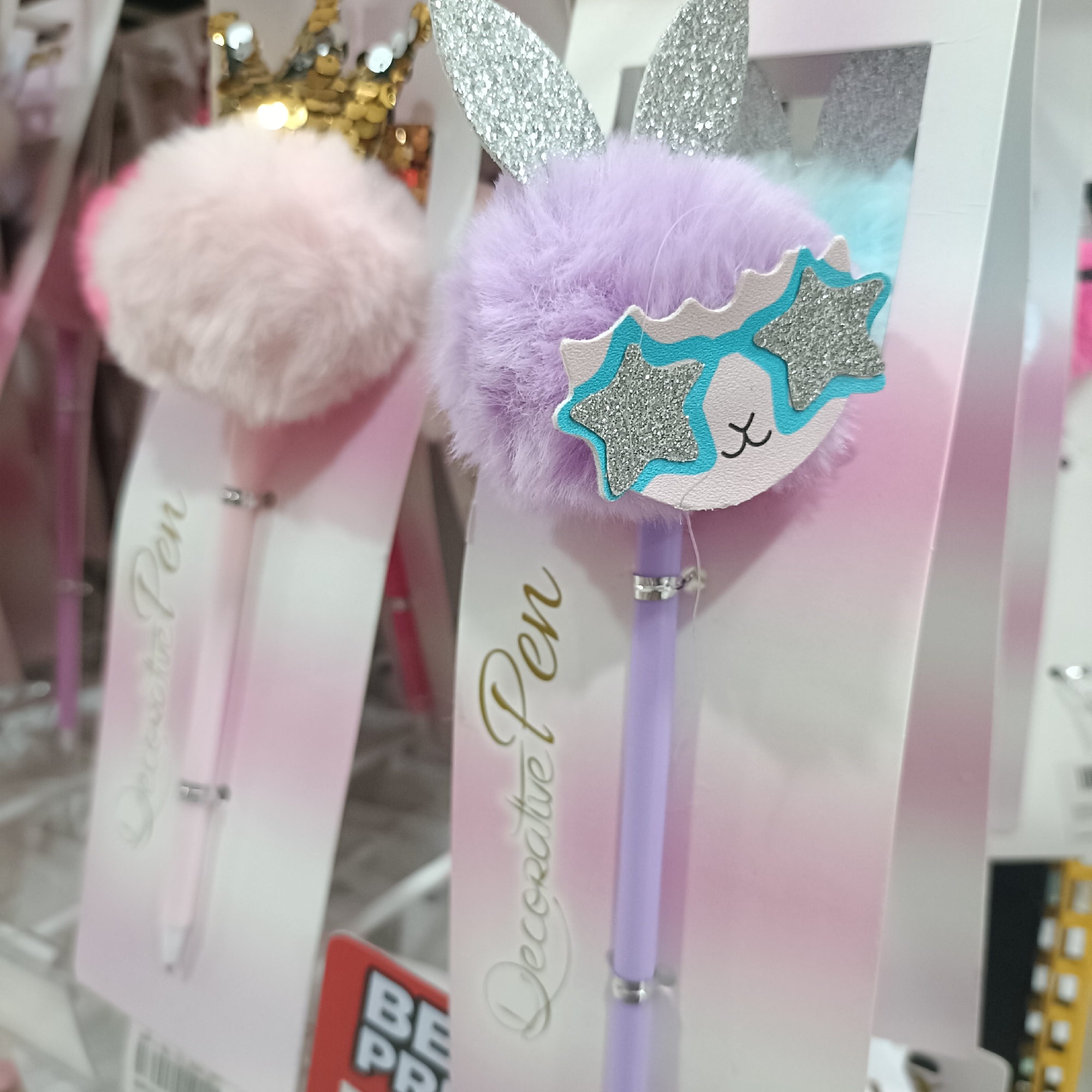 A close-up shot shows two "Decorative Pen" products in a retail setting. The pen on the left features a pink fluffy pom-pom with a gold crown detail, attached to a pink pen. The pen on the right has a lavender fluffy pom-pom with silver glitter bunny ears, adorned with a pair of cartoon-like sunglasses. The pens are mounted on packaging cards, one of which partially obscures a red and white sign ("BE PR") in the lower left corner. The scene takes place in a commercial environment in Madiun, Indonesia.