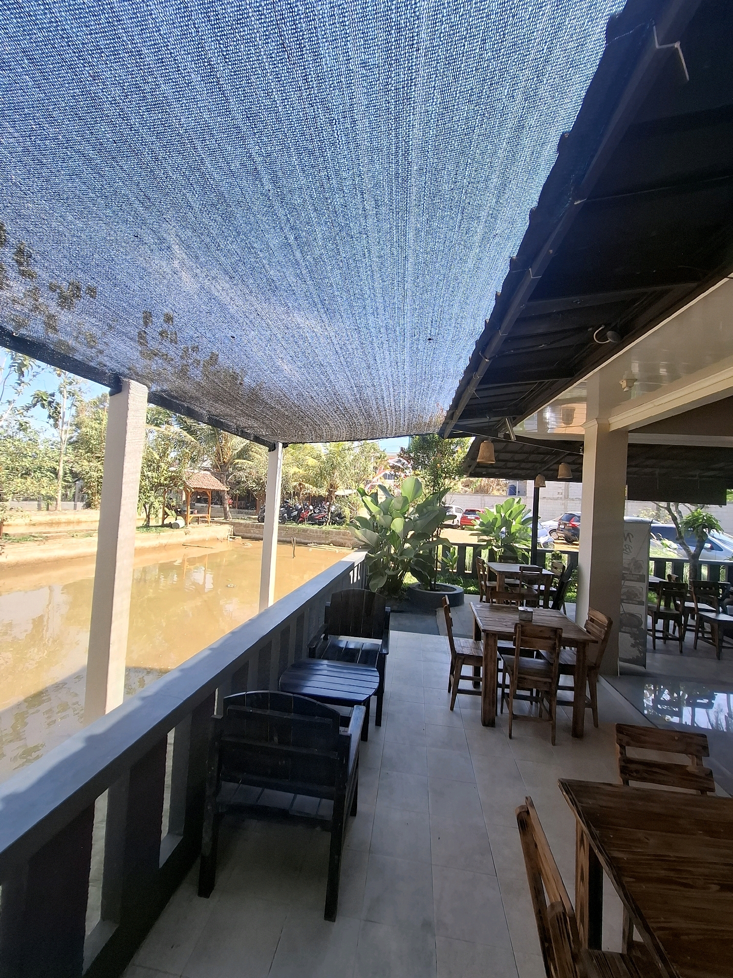 A shaded outdoor seating area overlooks a murky brown pond. The seating area is paved with light-colored tiles and furnished with several wooden tables and chairs. A dark blue mesh shade covers the area, supported by a black metal frame and white pillars. Beyond the railing of the seating area, the pond is visible, with trees and foliage surrounding it. In the background, a small wooden gazebo sits near the water's edge, and parked motorcycles are visible. The scene appears to be in Cibentang, Indonesia, likely a restaurant or cafe with an outdoor dining space. The lighting suggests it is daytime, with sunlight filtering through the mesh shade.