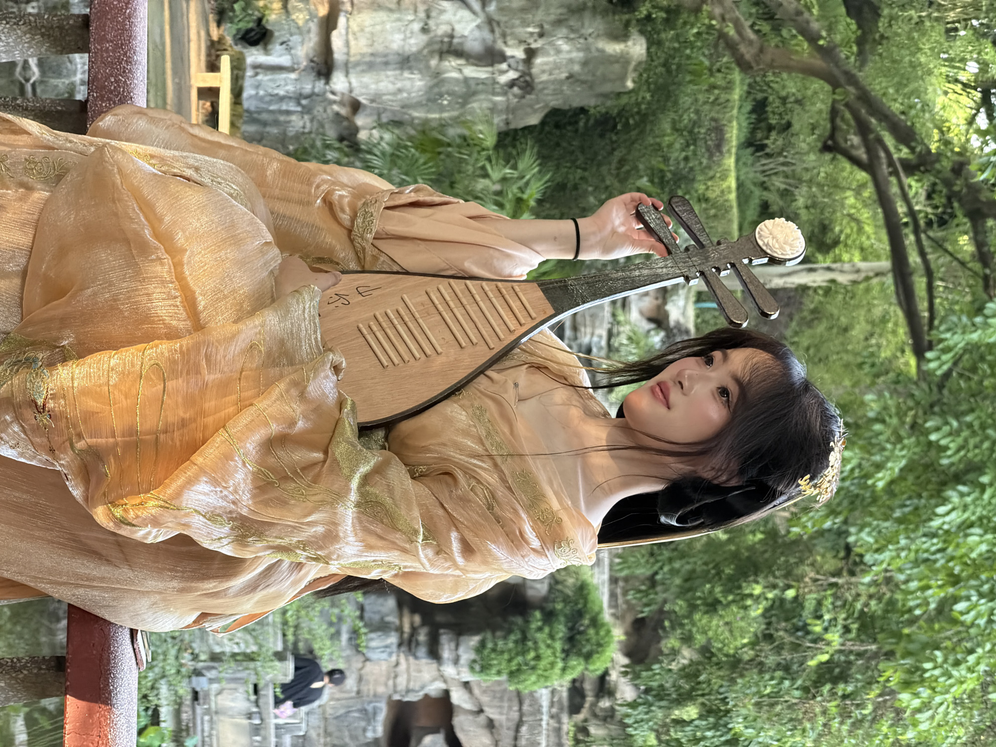In an outdoor garden setting, likely in Yuzhong District, China, a young woman is seated on a railing, holding a traditional Chinese lute. She is dressed in an elaborate, flowing peach-colored gown with golden embroidery. Her dark hair is styled with ornate gold ornaments. The lute, made of wood, has four strings and a decorative white embellishment at the top. The background features lush green foliage, rock formations, and water, suggesting a traditional Chinese garden or park. Sunlight filters through the trees, illuminating the scene. In the distance, a person in a black shirt is visible walking up stone steps. The overall impression is one of serene beauty and cultural representation.