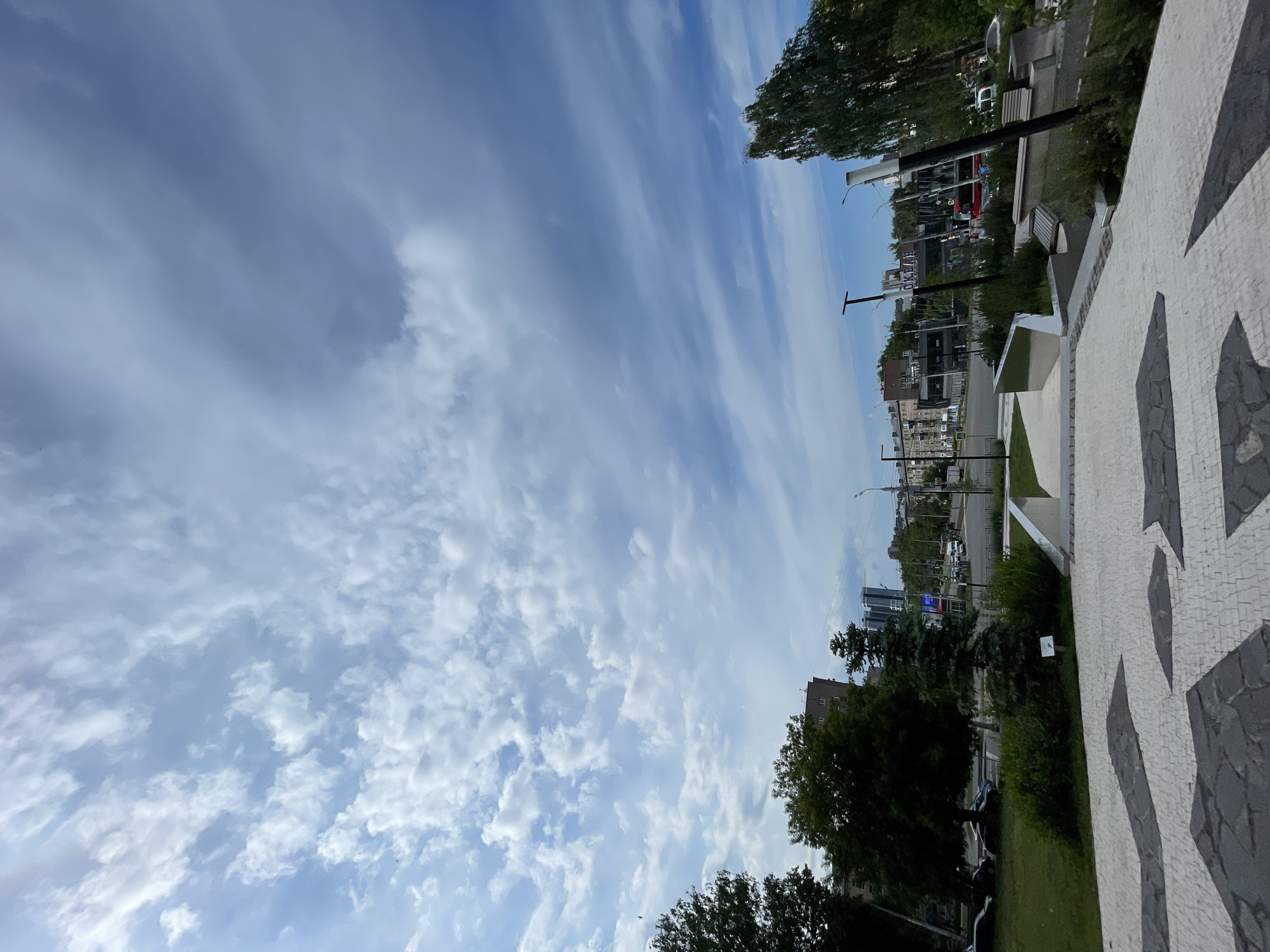 The image captures a wide urban landscape under a bright, dynamic sky. The upper two-thirds of the frame are filled with a light blue sky featuring numerous cloud formations. These clouds range from delicate, wispy streaks to more defined, scattered cumulus-like patches, creating a textured overhead vista.
In the foreground, on the right side of the image (when viewed horizontally), there is a distinctive paved area. This surface is adorned with a mosaic pattern primarily composed of small white tiles, accented by larger, irregular dark grey or black geometric shapes. Moving towards the midground, adjacent to this paved area, is a strip of green grass dotted with bushes and several trees displaying dense green foliage.
The background reveals a bustling urban environment. A street runs parallel to the foreground, lined with various buildings, including multi-story residential structures and more modern, possibly commercial buildings with reflective surfaces. Streetlights are visible along the street, and there are hints of parked vehicles or light traffic. The setting appears to be a city park or a wide promenade alongside a main thoroughfare.
The scene is illuminated by daylight, suggesting a clear or partly cloudy day. The overall atmosphere is calm and open. There are no identifiable people or explicit activities taking place. No discernible text is visible on any signs or buildings within the frame.