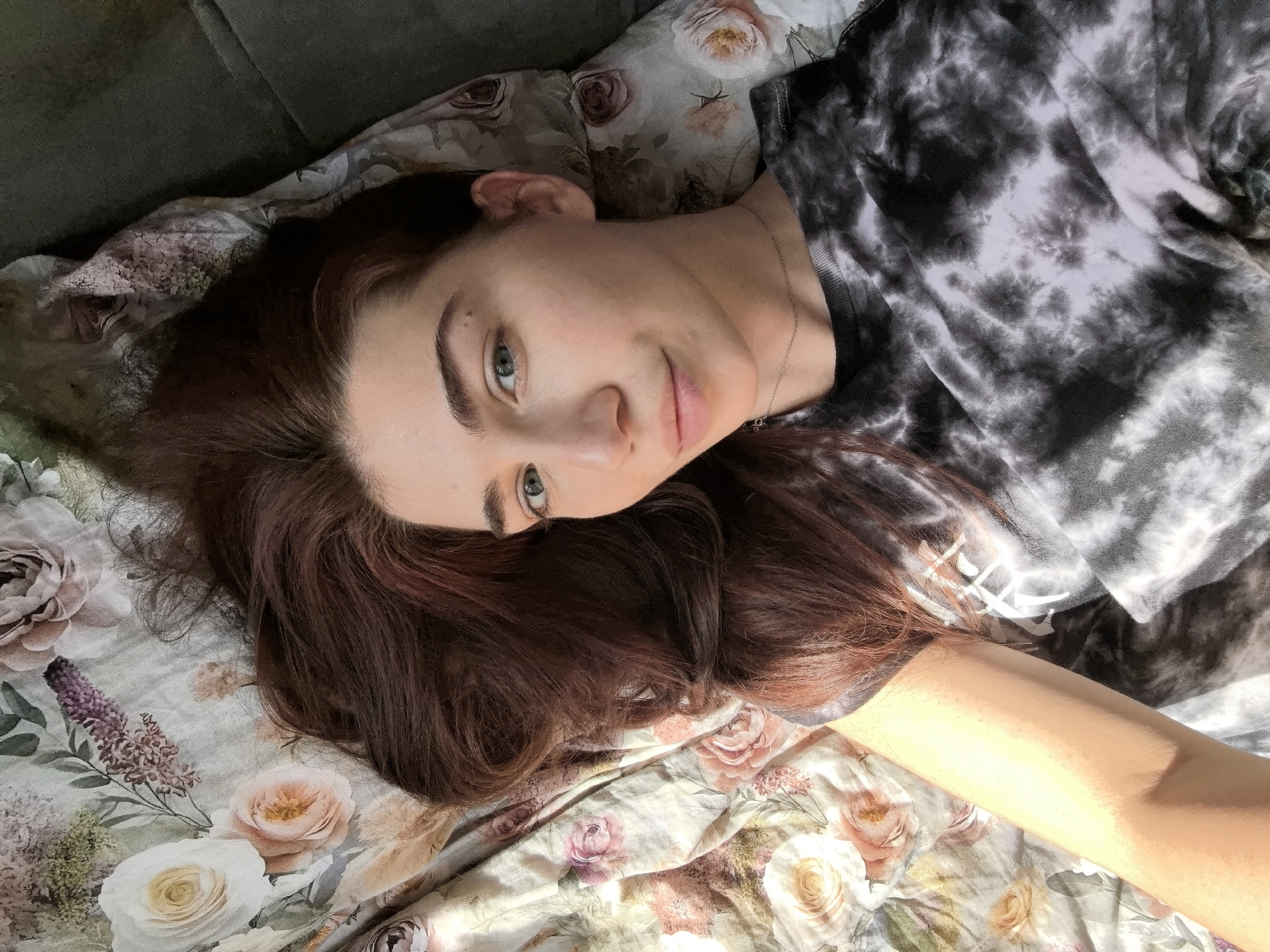 A woman is lying down, presumably indoors, on a floral-patterned surface, possibly a bed or sofa. She has fair skin, auburn hair, and light-colored eyes. She is wearing a gray and black tie-dye shirt. Her arm extends towards the bottom right of the frame. The location of Stara Zagora, Bulgaria, cannot be determined from the visual information provided in the image.