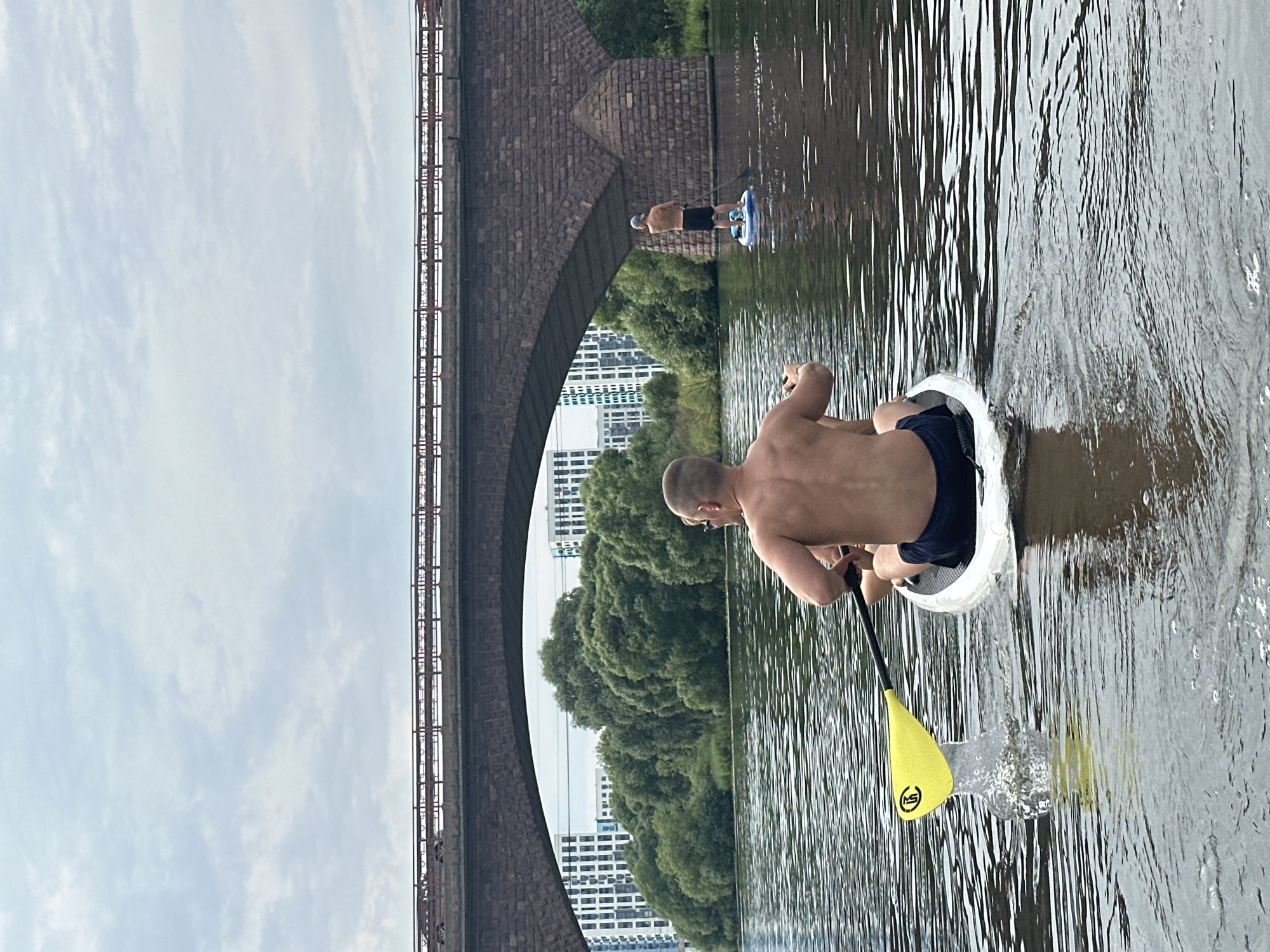 Two shirtless men are paddleboarding on a calm river. The man in the foreground is seated on his paddleboard, facing away from the camera, and is in the process of paddling, with his yellow paddle creating a splash in the water. The second man is standing on his paddleboard further down the river, also facing away from the camera, and is paddling. Both men are wearing dark-colored shorts.
Behind the men, a large stone bridge with arched openings spans the river. Through the arches, a dense line of green trees is visible, interspersed with modern white buildings. The sky is overcast with clouds. The scene appears to be set in the city of Yam, Russia, suggested by the presence of a river, bridge, and urban development.