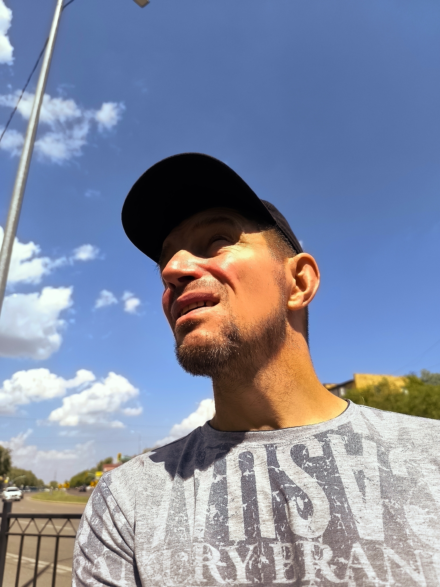 A man wearing a black baseball cap and a gray t-shirt with distressed lettering stands outdoors under a bright blue sky with scattered white clouds. He is looking upwards and to his left, with sunlight illuminating his face, creating shadows. The background shows a street with trees, buildings, a fence, and a few cars. The image is a low-angle shot, focusing on the man's face and upper body.