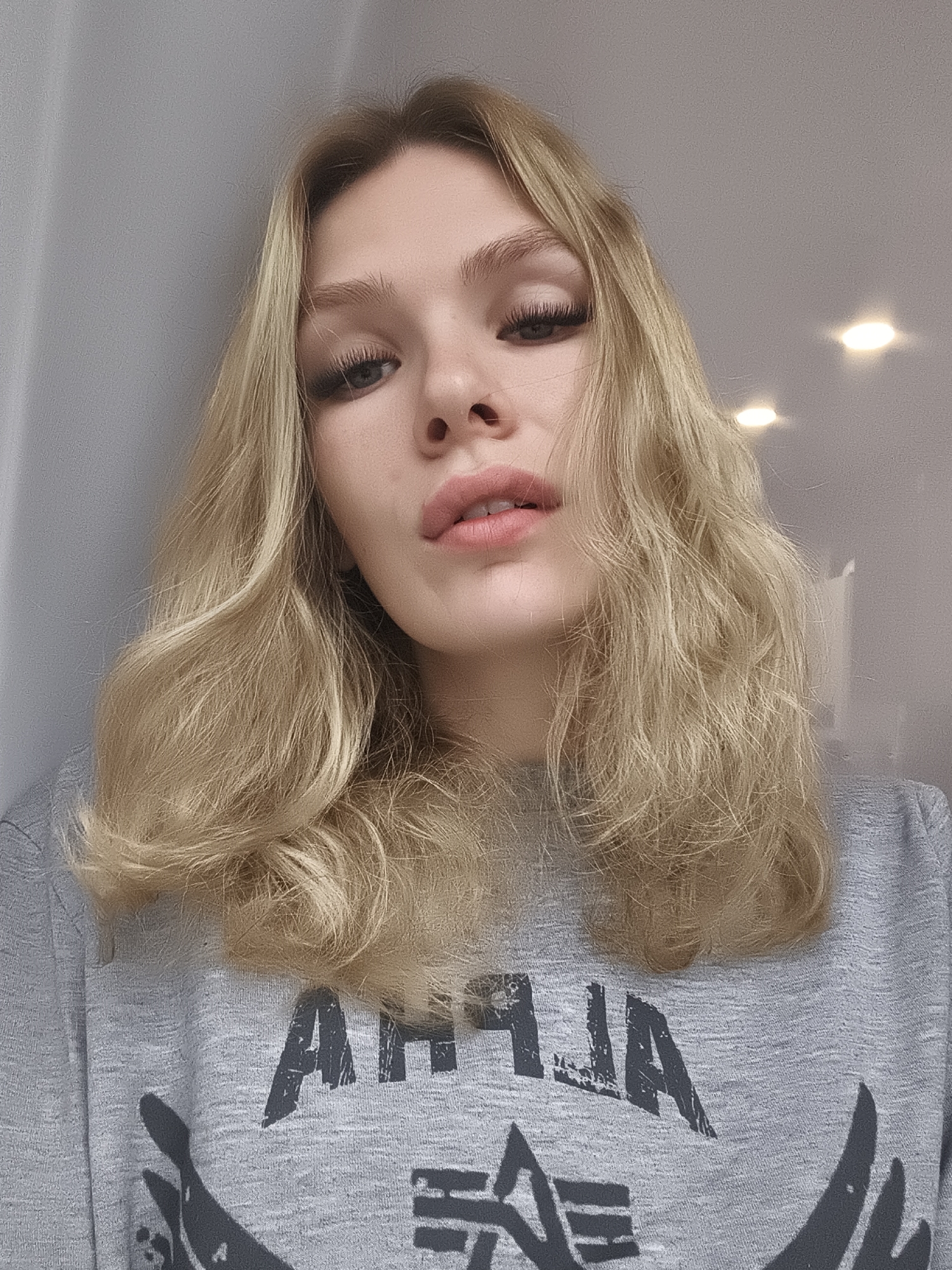 A fair-skinned young woman with blonde wavy hair is centered in the frame, looking slightly down at the camera. She has prominent cheekbones, full lips, and visible eyelashes. She's wearing a gray sweatshirt with a large, dark graphic print that appears to have the word "ALPHA" and wing-like images. The background includes a plain, light gray wall on the left and a ceiling with two recessed lights on the right. The scene appears to be indoors. There is no specific evidence to link the image to the city of Русский Вожой, Russia.