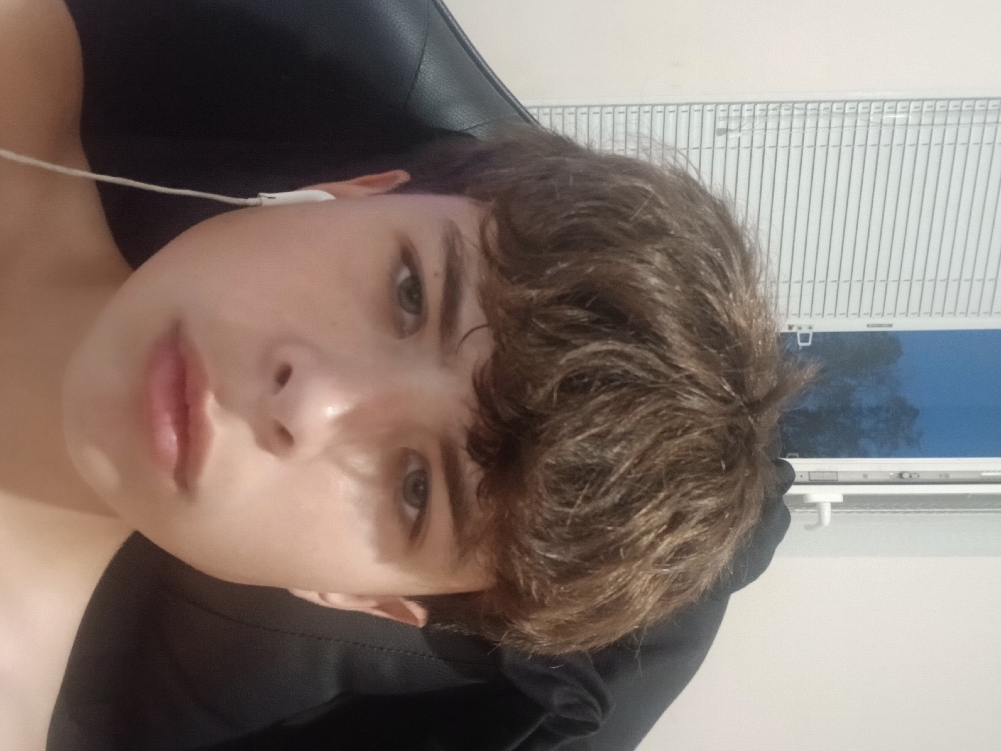 A young male with brown hair and light skin is looking directly at the camera. He is wearing white earbuds. He is seated in a black chair. Behind him, there is a closed white window with blinds, and to the right of the window, there is a wall. The image appears to be taken indoors.