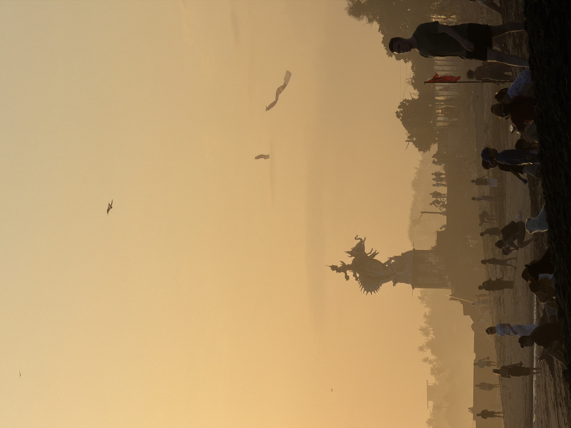 A silhouetted scene unfolds at sunset on a beach in North Kuta, Indonesia. A large, elaborate dragon-like statue, perched atop a multi-tiered structure, dominates the mid-ground, silhouetted against the hazy, orange sky. Numerous people are scattered across the sandy beach, some walking, others sitting or interacting in small groups. In the distance, a line of trees and buildings create a hazy backdrop. Two long, ribbon-like kites are visible in the sky, along with a few small birds. The overall impression is one of a tranquil evening gathering on a tropical shore.