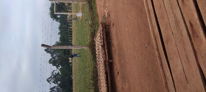 The image shows a scene in Lwandeti, Kenya, located at latitude 0.563 and longitude 34.806. The scene is a dirt field with a wooden structure in the foreground. There is a wooden bench in the middle ground. In the background, there is a green field, a fence, and trees. Several people are visible in the distance. The people are walking and playing in the field. The scene is taken from a slightly elevated vantage point, looking towards the field. The weather is cloudy.