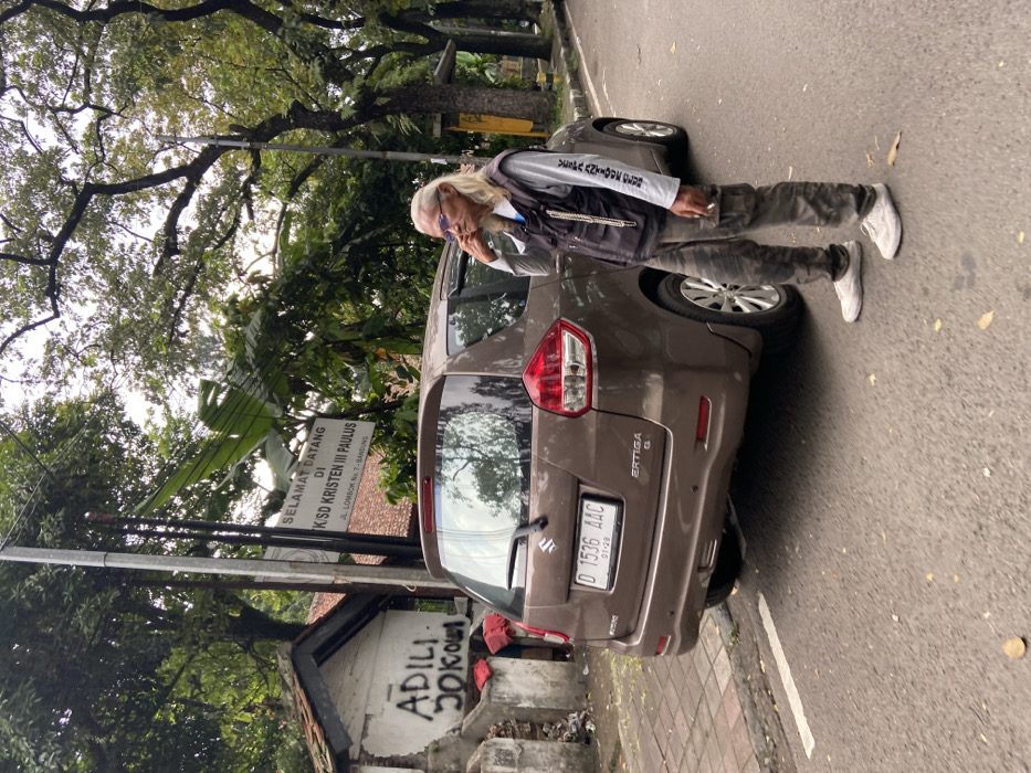 The image shows a man standing next to a brown Suzuki Ertiga car parked on a paved street in front of a building in Bandung, Indonesia, located at latitude: -6.911, longitude: 107.618. The man is wearing a grey t-shirt with black writing on it, a black vest with silver chains hanging from the zipper, and camo cargo pants. He is holding a cigarette in his right hand and looking to the side. The car has a license plate that says "D 1536 AAC". The building in the background has a sign that reads "Selamat Datang Di KSD Kristen III Paulus JL. Lombok No. 7 - Bandung". There is graffiti on the wall of the building that reads "ADILI DOKOL". The street is paved and has a white line marking the edge of the road. There are trees and plants in the background. The sky is overcast.