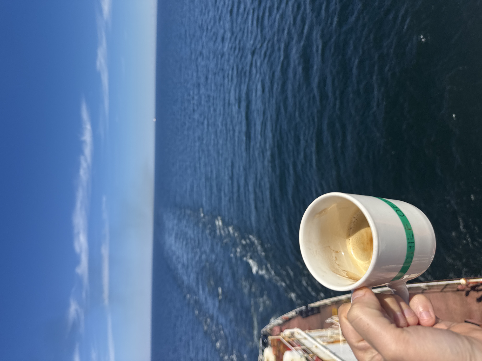 A person's hand holds a white mug with a green band, filled with coffee, against the backdrop of the ocean. The mug has the words "CHIEF OF" visible on the green band. The background shows the dark blue ocean with a wake trailing behind a ship on the left side of the frame. The sky is clear and blue with a few wispy clouds. A faint smoke plume is visible on the horizon. The scene appears to be on a ship at sea.
